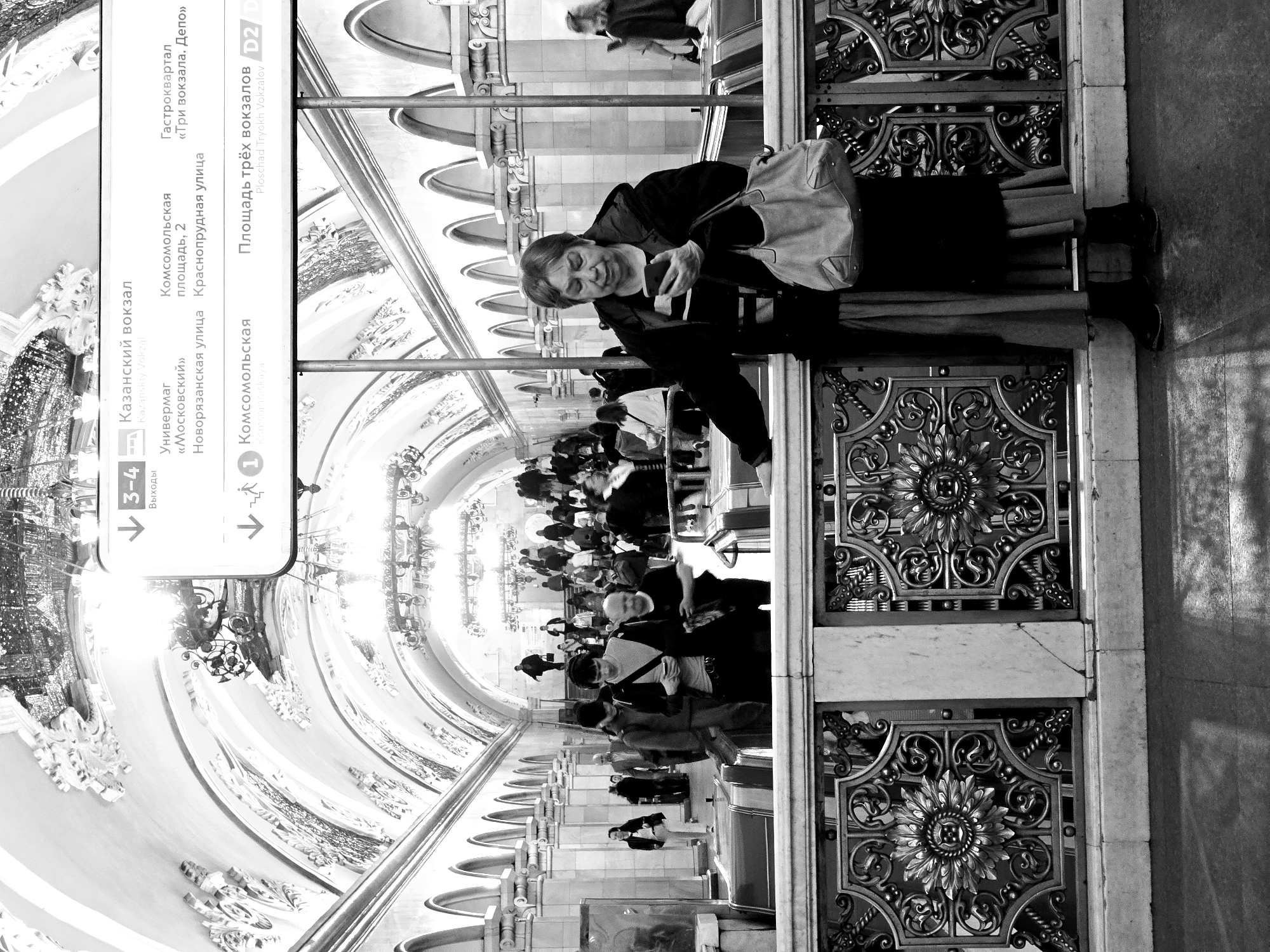 The image, shot in black and white, appears to depict an indoor scene, possibly a train or subway station in Moscow, Russia, given the signage and architecture. A woman is holding a phone, seemingly taking a picture or using it while standing near an ornate metallic barrier. The backdrop shows an arched corridor with intricate artwork along the walls. People are walking in the background. Signs in Russian are visible, indicating directions to various locations such as Kazansky Vokzal (Kazan Railway Station), Komsomolskaya, and "Three Vokzals Square."