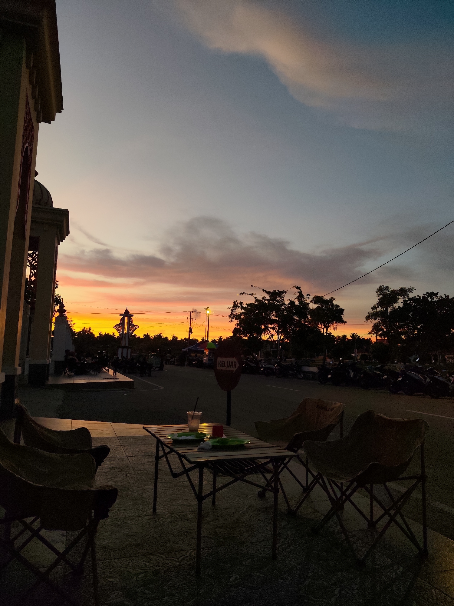The image captures a serene sunset scene in Meunasahaluedrienlhoksukon Barat, Indonesia. The sky is ablaze with vibrant hues of orange, yellow, and pink, indicating the golden hour. In the foreground, a set of foldable chairs and a table are arranged on a tiled patio, suggesting a casual outdoor dining or relaxation area. Two chairs are prominently visible on the left, and another two are placed around the table, which holds a drink and some plates.
Beyond the seating area, a road runs horizontally, lined with parked motorcycles. Trees with dark foliage create a silhouette against the colorful sky, adding depth to the scene. To the left, a portion of a building with intricate architectural details, possibly a mosque given the location context, is visible. People are seen seated and standing near this building, engaged in quiet conversation or observation of the sunset. A prominent decorative pillar stands near the building.
Further down the road, stalls and lights are faintly visible, hinting at a bustling marketplace or gathering spot that comes alive in the evening. A red circular sign with white text that reads "KELUAR" (meaning "Exit" in Malay and Indonesian) is positioned on the right side of the road. The overall mood is peaceful and tranquil, evoking a sense of calm as the day transitions into night, enhanced by the warm glow of the setting sun. The weather appears clear, with scattered clouds catching the sunset colors.