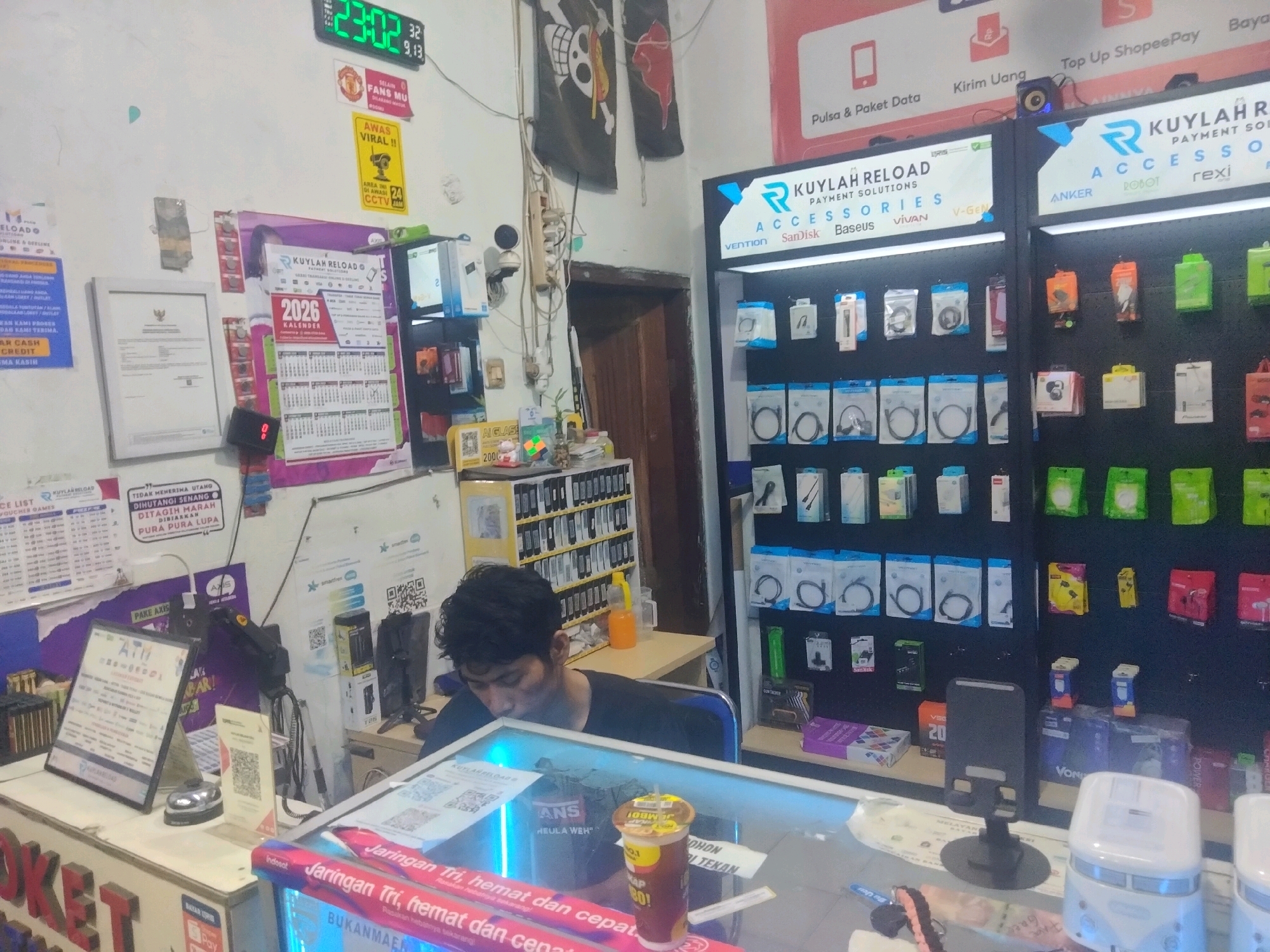 The image depicts the interior of a small retail store, likely a mobile phone and accessories shop. The scene is filled with various electronic accessories displayed on shelves, including cables, chargers, and earphones. Prominent signage indicates "KUYL AHRELOAD PAYMENT SOLUTIONS ACCESSORIES". A digital clock on the wall displays "23:02". A young man with dark hair is seated behind a counter, appearing to be working or attending to customers, though no direct interaction is visible. The environment appears somewhat cluttered, with posters, calendars, and other informational materials displayed on the walls. The presence of multiple QR codes suggests a focus on digital payments and services. The lighting is functional, typical of a commercial establishment. The overall impression is of a busy, independently-owned store operating in a local community.