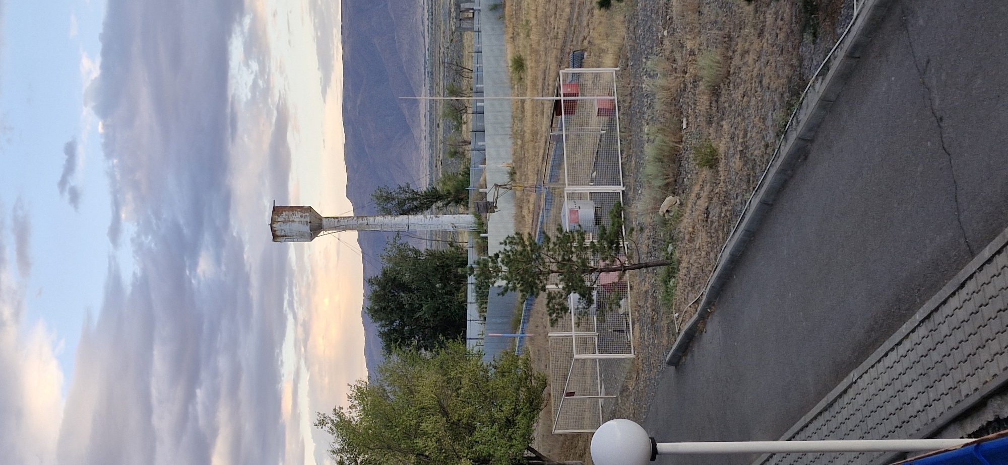A low-angle, slightly elevated outdoor shot captures a scene in Usharal, Kazakhstan, featuring a prominent, weathered water tower against a backdrop of muted hills and a cloudy sky. The water tower, tall and slender, shows signs of rust and wear, particularly at its top. In the foreground, a paved pathway runs diagonally from the bottom left, bordered by a textured walkway and a curb. To the right of the pathway, a white lamppost with a spherical lamp is visible.
Beyond the immediate foreground, a fenced-off area contains a cylindrical structure with red accents, likely a pump or storage tank, and a small pine tree stands within the enclosure. A chain-link fence surrounds this area, extending into the middle ground. Further back, a row of trees, predominantly green, flanks the water tower and the fenced compound. A long, grey metal fence stretches across the mid-ground, separating the nearer elements from the drier, more sparse vegetation and distant, rolling hills that dominate the horizon. The sky above is a mixture of light blue and grey clouds, with a hint of soft sunlight breaking through on the left side, suggesting either sunrise or sunset. No people or animals are visible in the frame. The overall impression is one of a functional, perhaps industrial or infrastructural, site within a semi-arid landscape.
