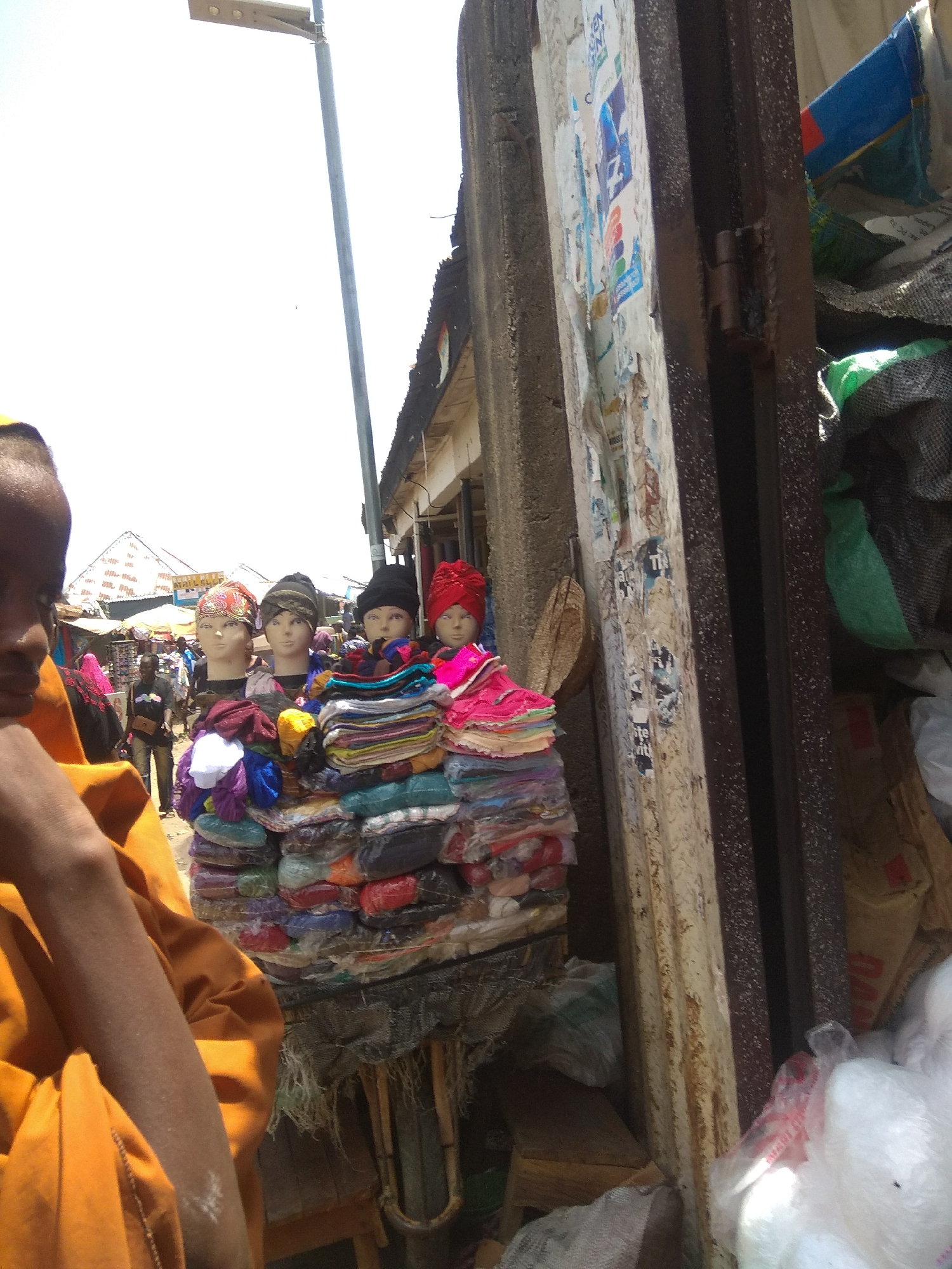 The image depicts a street market scene in Jalingo, Nigeria. In the foreground, a person wearing an orange garment is partially visible on the left. In the center, a stall displays various colorful fabrics and headwear on mannequins. The mannequins are adorned with turbans and headwraps. Behind the stall, more market stalls and people are visible. On the right side of the image, a building facade with peeling posters and a metal door is present. The sky is bright and overcast.