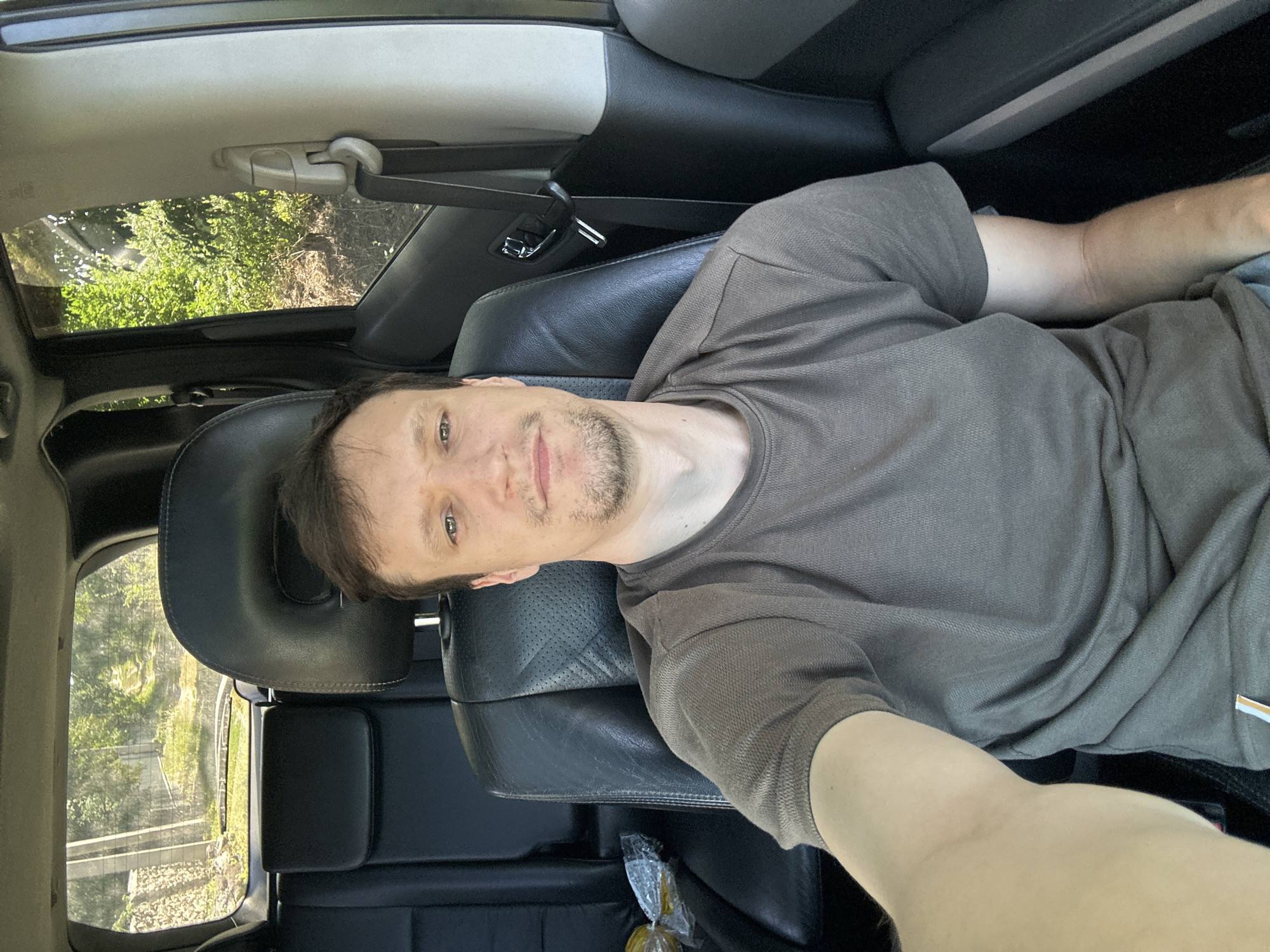 A young adult male with light brown hair and a light beard is seated in the driver's seat of a car, taking a selfie. He is wearing a brown t-shirt. The car's interior is visible, including black leather seats and part of the dashboard. Outside the car, through the windows, a rural landscape with green trees and a dirt road can be seen. The image was captured on a sunny day. The specific location within Makiv, Ukraine cannot be determined from the visual information alone.