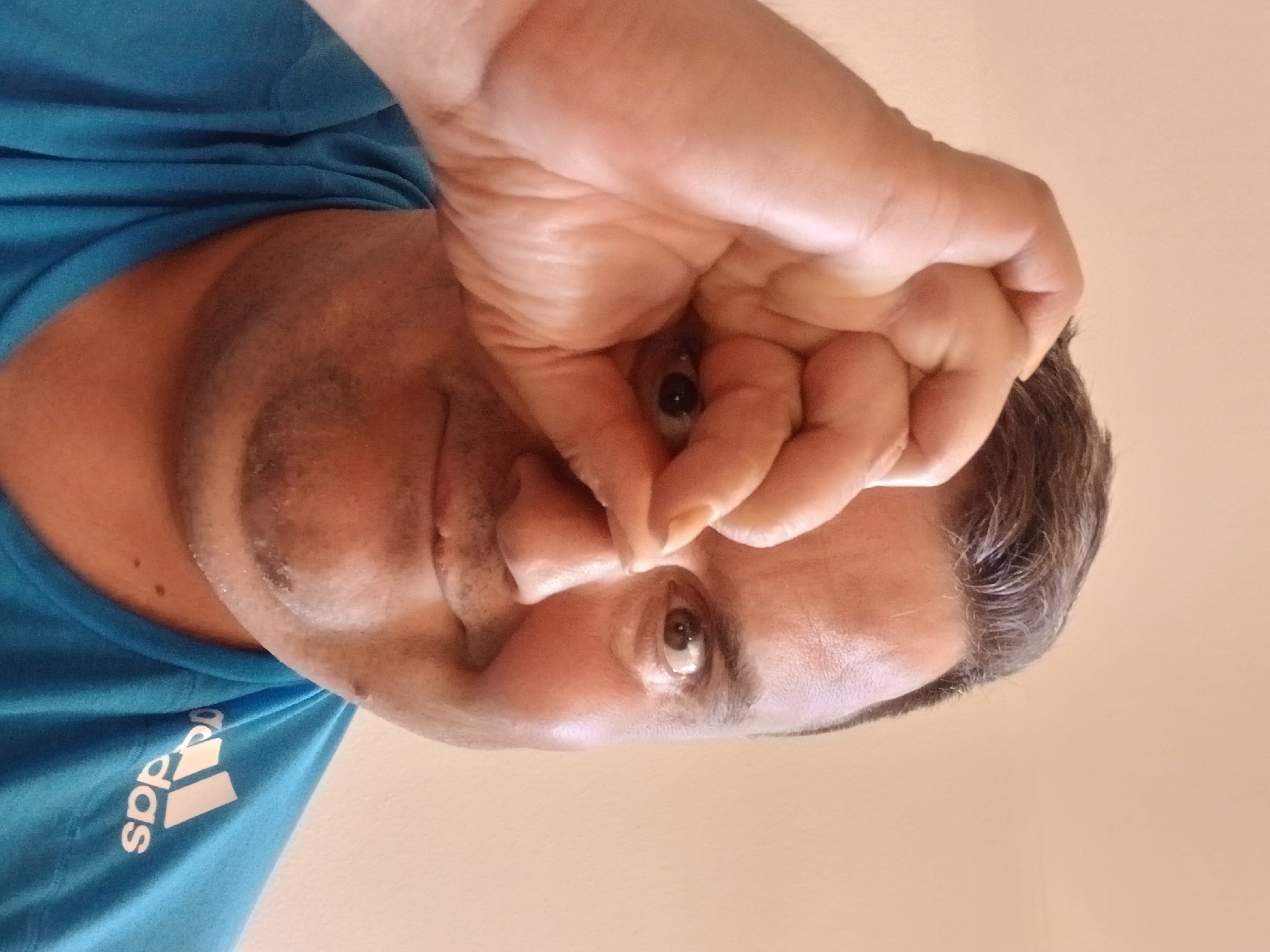 A medium shot captures a man from the chest up, looking directly at the camera. He has fair skin, dark hair, and is wearing a blue t-shirt with the Adidas logo on the right shoulder. His right hand is raised to his face, with his thumb and index finger forming a circle around his right eye. His left eye is visible, and he has a slight smile. The background is a plain, light-colored wall. The image appears to be taken indoors with natural light. There is no identifiable information about the location in Pfarrkirchen, Germany, or the man's identity.