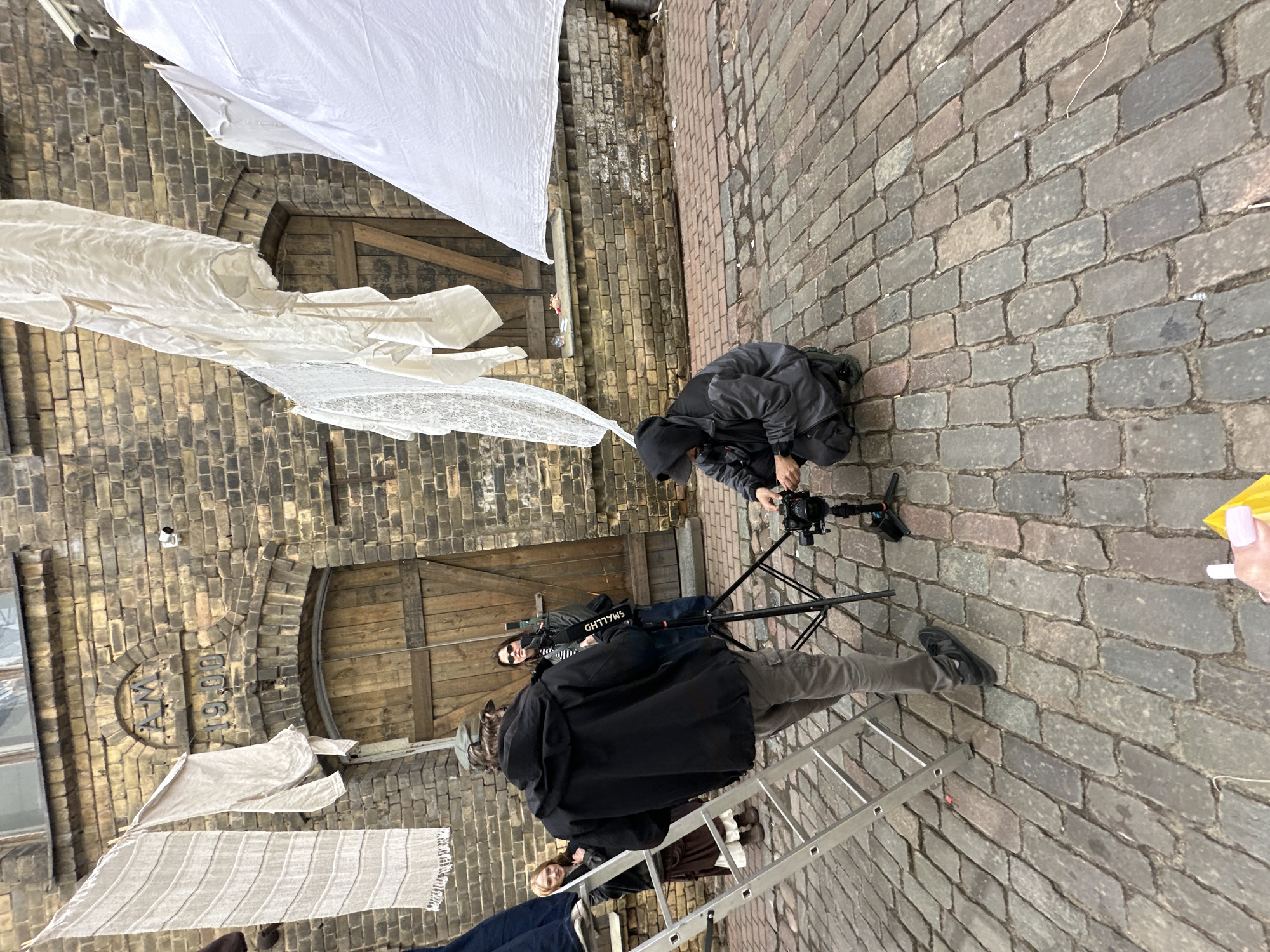 The image captures a scene in Riga, Latvia. Several individuals are present in a narrow cobblestone alleyway surrounded by aged brick buildings with large wooden doors. White fabric is suspended overhead, resembling laundry or decorative displays. One person kneels near a camera on a tripod, likely filming or photographing. Additional people are visible: one stands wearing sunglasses, and another stands further back. A metal ladder is present, and a hand holding a yellow object is visible in the lower right corner. The alleyway is lined with worn brick buildings, one of which has "A.M. 1900" carved into the brickwork above a wooden door.