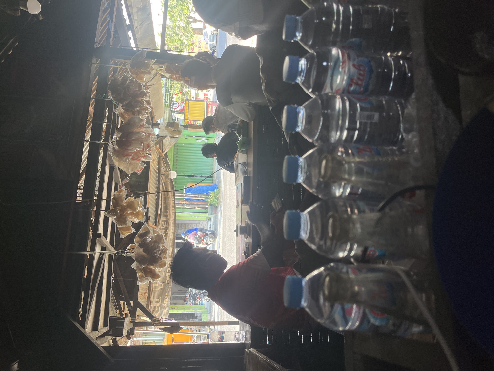 The image depicts a street-side food stall in Dusun Metatu, Indonesia. The stall is open-air, with a corrugated metal roof supported by wooden beams. Bags of dried goods, likely snacks or ingredients, are hung from the ceiling. In the foreground, several plastic bottles of water are arranged on a surface, some partially obscured.
Inside the stall, a man wearing a red shirt is seated at a counter, looking down at something in his hands, possibly a phone. He appears to be interacting with the counter which has various items on it, including a small cup. Behind him, other customers are visible, some seated and facing away from the camera, suggesting they are eating or drinking.
The background shows a street scene with vehicles, including motorcycles and a car, and buildings. The lighting suggests it is daytime, with sunlight filtering into the stall. The overall impression is of a casual, local eatery.