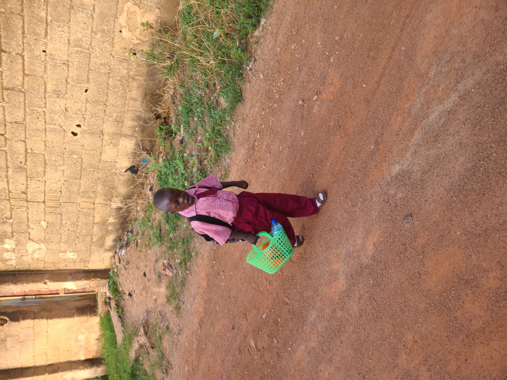 A young person in a school uniform is walking along a dirt road in Jos, Nigeria. The individual wears a red and white checkered shirt and maroon pants with dark shoes. They carry a lime green plastic basket containing a water bottle. The backdrop features a weathered brick building, with green vegetation growing along the roadside. The ground is composed of reddish-brown dirt, and the overall scene has a somewhat rural or undeveloped appearance.