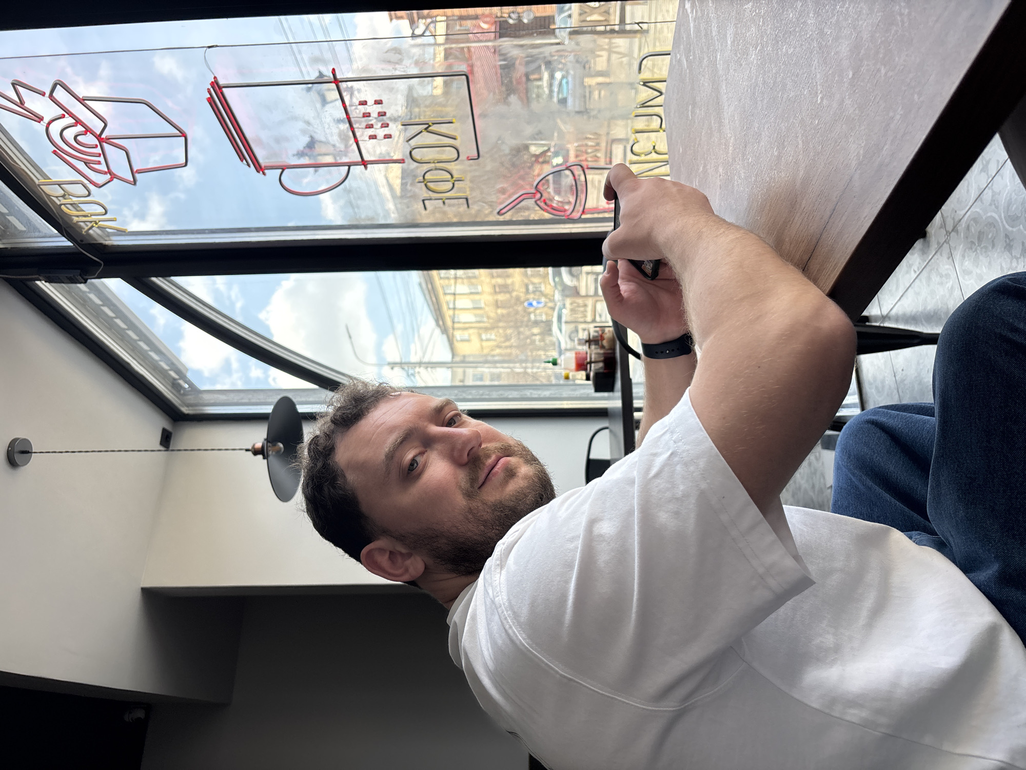 A man with a beard and dark, curly hair, wearing a white t-shirt and blue jeans, is seated at a wooden table in what appears to be a cafe in Krasnoyarsk, Russia. He is looking directly at the camera with a neutral expression, while holding a smartphone with both hands, angled away as if taking a photo or video. A dark-colored smartwatch is visible on his left wrist.
The setting is bright, with a large window behind him showcasing a clear, cloudy blue sky and reflections of urban buildings outside. Adhered to the window pane are neon-style signs. One prominent sign clearly spells "КОФЕ" (KOFE), which means "COFFEE" in Russian, next to an illustration resembling a coffee press. Another partial word, possibly "BOK," is visible on the left, and "ВКУМ" (VKUM) is reflected on the right.
An industrial-style black ceiling light fixture hangs from the ceiling to the left. The floor beneath the table has a patterned tile. The overall impression is a daytime scene in a modern cafe environment.