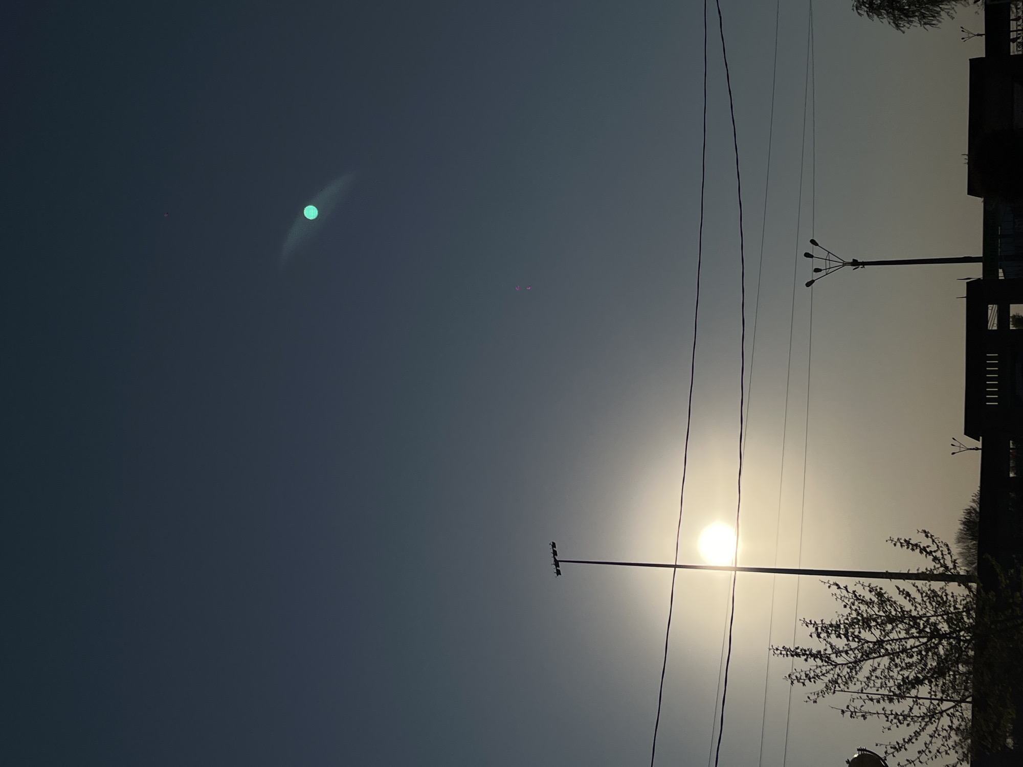 The image shows a twilight scene in Novopokrovskaya, Russia. Overhead power lines are visible, along with silhouetted buildings and trees on the right side. A street light pole is in the foreground. The sun is obscured by haze, positioned in the middle. In the upper left portion of the image are a small red dot, and green flare. The sky transitions from dark blue to lighter yellow in the lower right portion of the picture.