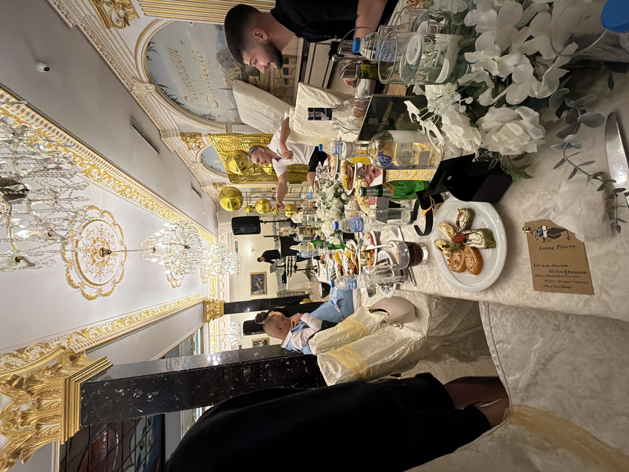 The image depicts a lavish indoor event, likely a banquet or celebration, with opulent decor characterized by ornate gold accents on the ceiling and pillars, and elaborate crystal chandeliers. A long, formally set table dominates the foreground, laden with food, drinks, and floral arrangements. In the mid-ground, several individuals are seated at the table. One man, wearing a light blue suit and white shirt, appears to be speaking. To his right, another man in a black shirt is seated, looking towards the left. In the background, a DJ setup with speakers is visible, suggesting entertainment. The overall ambiance suggests a festive and upscale gathering.