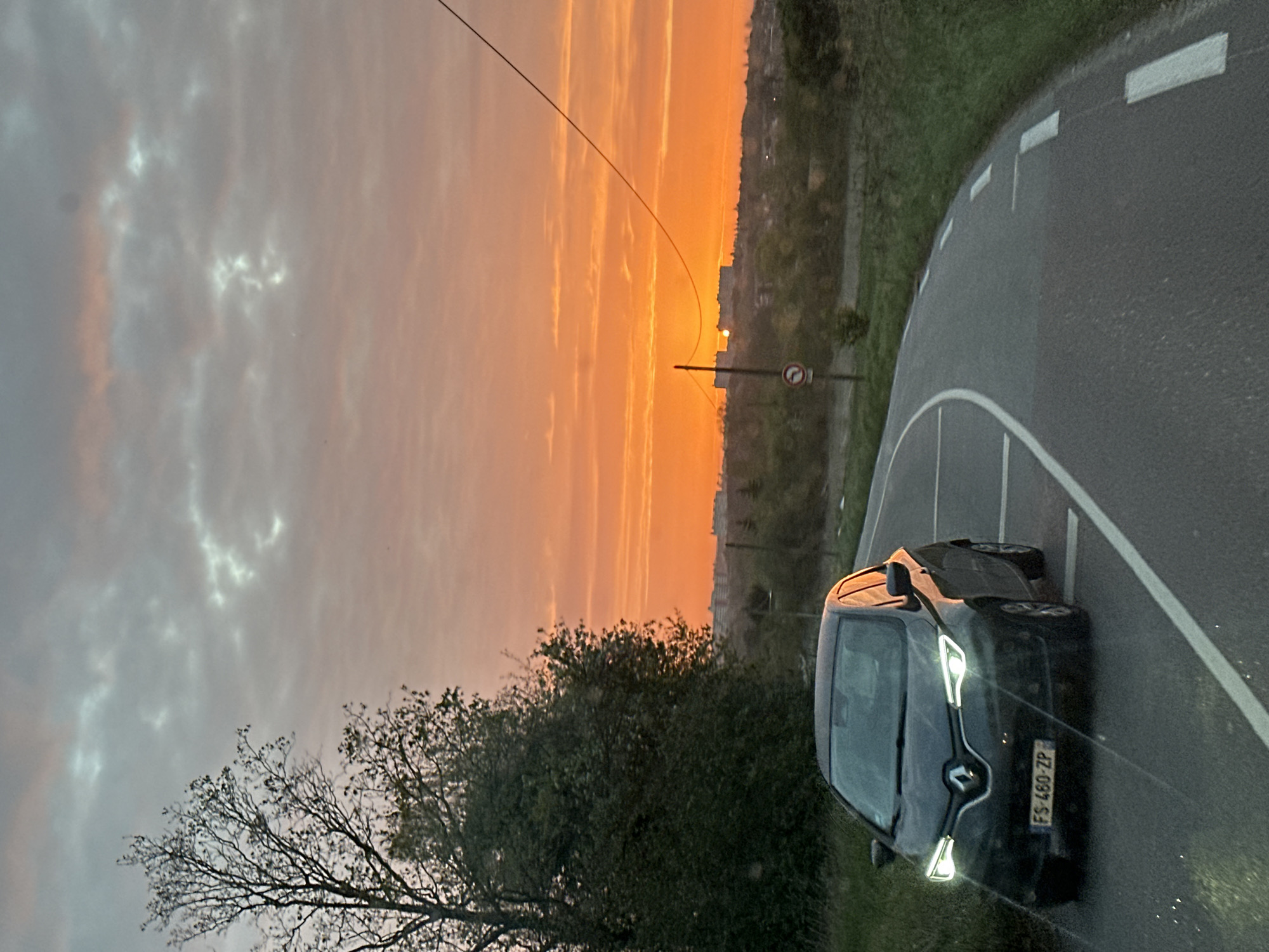 The image shows a dark-colored car on a road in Saulx-les-Chartreux, France. The road curves to the right, with white lines marking the lanes. There is a bush or tree along the side of the road and buildings in the distance, silhouetted against a bright orange sky. The sky appears to be at sunset or sunrise. There is also a no return sign visible in the upper right of the image.