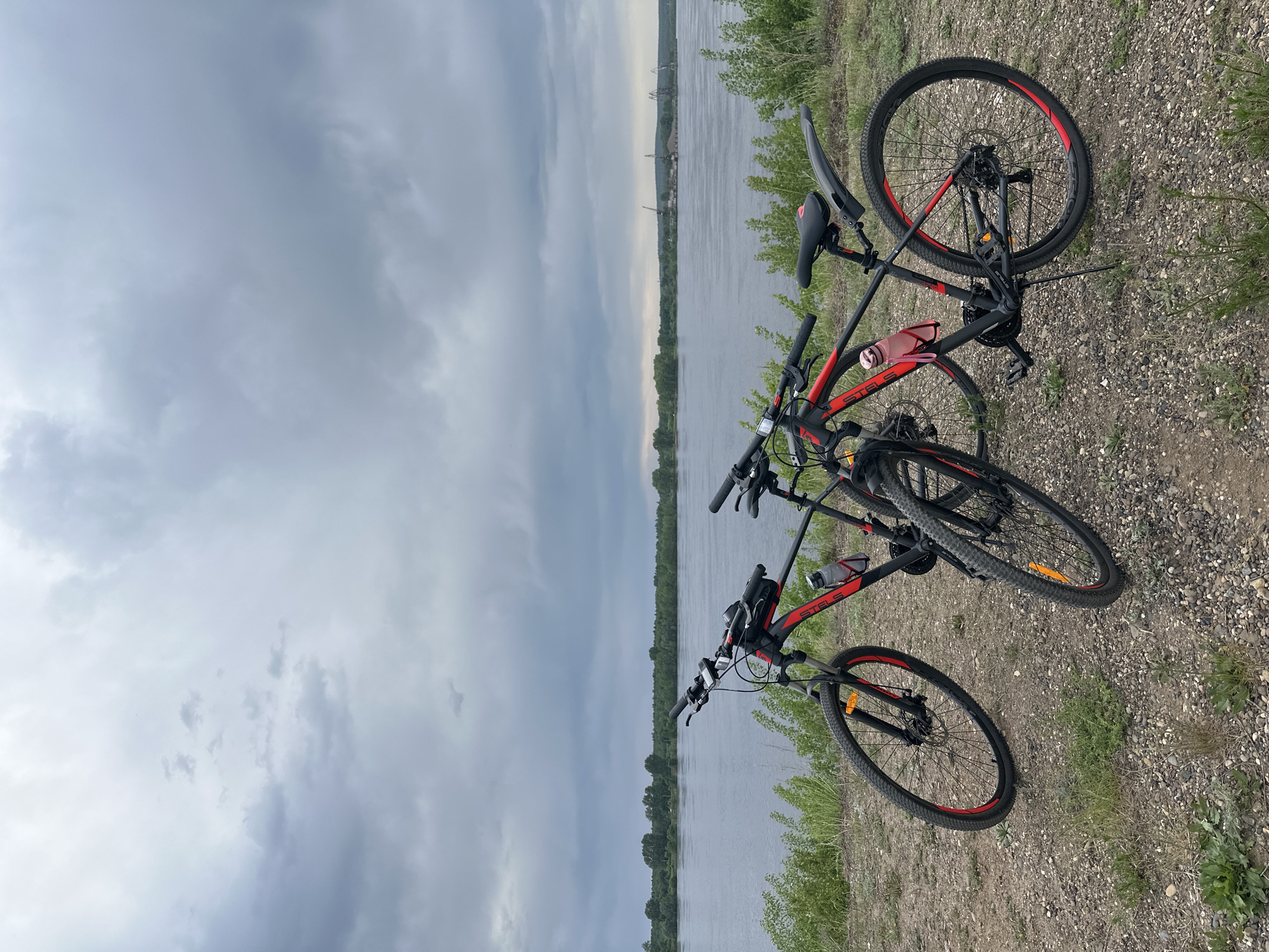 Two Stels mountain bikes are laying on their sides on a gravel and grassy bank. Behind the bikes is a wide river, bordered by trees. On the far side of the river, indistinct buildings or structures are visible in the distance. The sky is overcast.
Based on the presence of "STELS" mountain bikes and the river scene, this image is likely taken in or around the city of Chaikovsky, Russia.