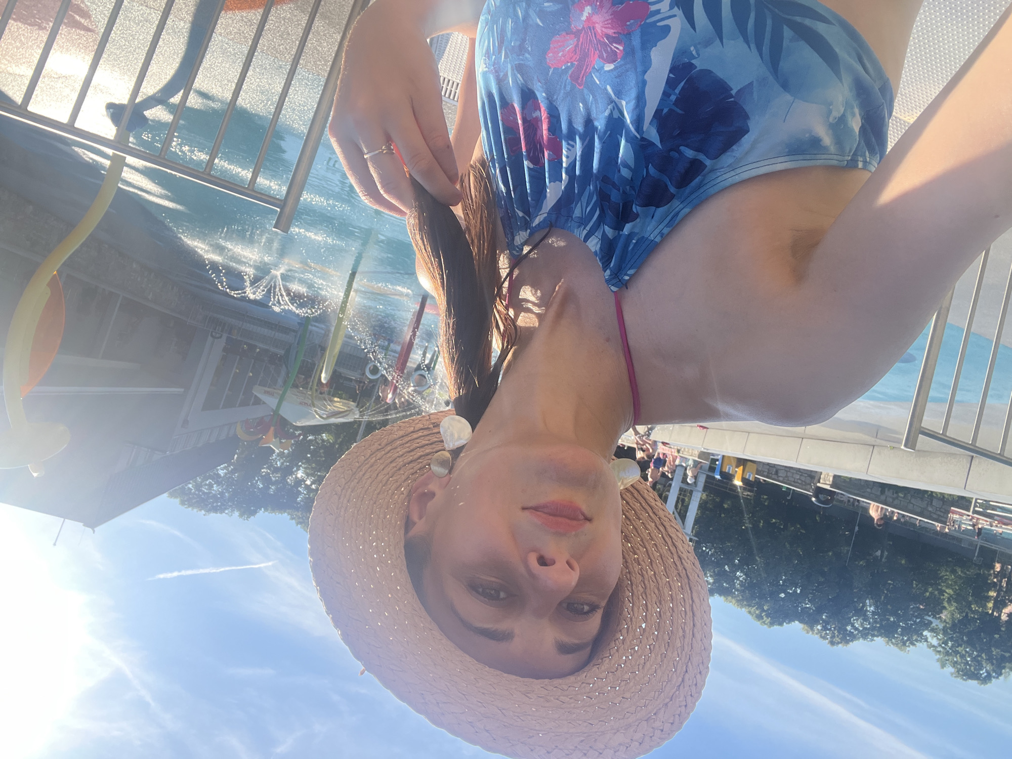 A woman is taking a selfie in a pool area. She is wearing a blue and white floral swimsuit and a straw hat. She has long brown hair and is wearing large, white, shell-like earrings. The sun is shining brightly, casting a warm glow on the scene, suggesting it is daytime. In the background, there is a swimming pool with water jets and a metal railing. There are also trees and what appear to be buildings visible beyond the pool area. Some people can be seen in the background, near the pool. The woman's expression is neutral. The image is oriented sideways.