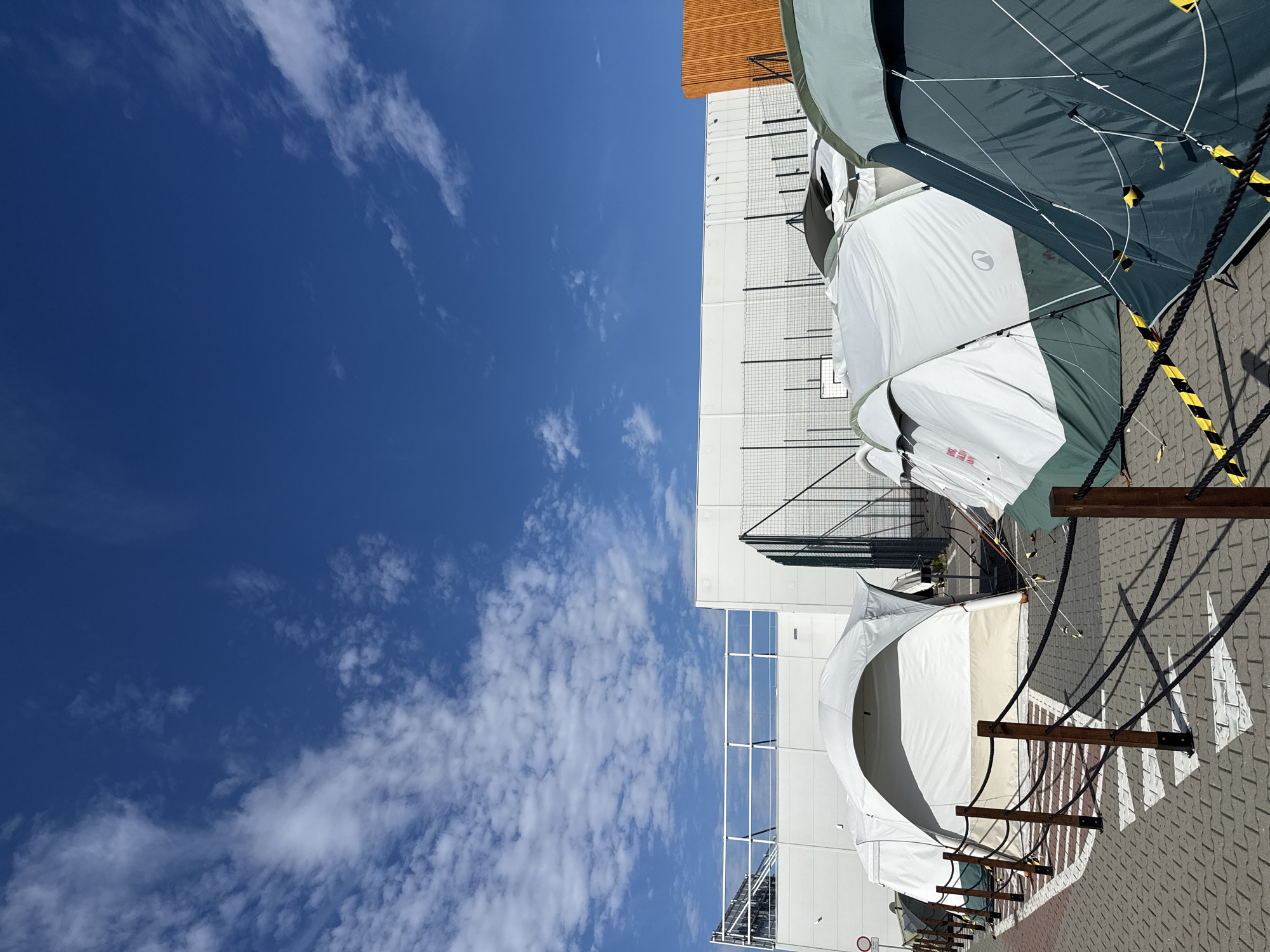 The image depicts an outdoor scene in what appears to be a temporary encampment, likely in Bielany Wrocławskie, Poland. Several large tents are set up in a row on a paved surface, possibly a parking lot or an open area adjacent to a building. The tents are of various sizes and colors, including white, green, and a combination of white and beige. Some tents are open, revealing their interior structures, while others are closed.
In the background, a modern, large building with a white facade and an orange-tiled section is visible. A white railing or fence runs along a raised platform in front of the building. A wire mesh fence is also present behind the tents. The ground is paved with interlocking bricks, and a temporary rope barrier with wooden posts separates the tents from a walkway. Some white markings, possibly directional arrows, are painted on the pavement.
The sky above is a bright blue with scattered white clouds. The overall impression is one of an organized, temporary shelter setup under a clear sky, with commercial or industrial buildings forming the backdrop. There are no people visible in the frame.
