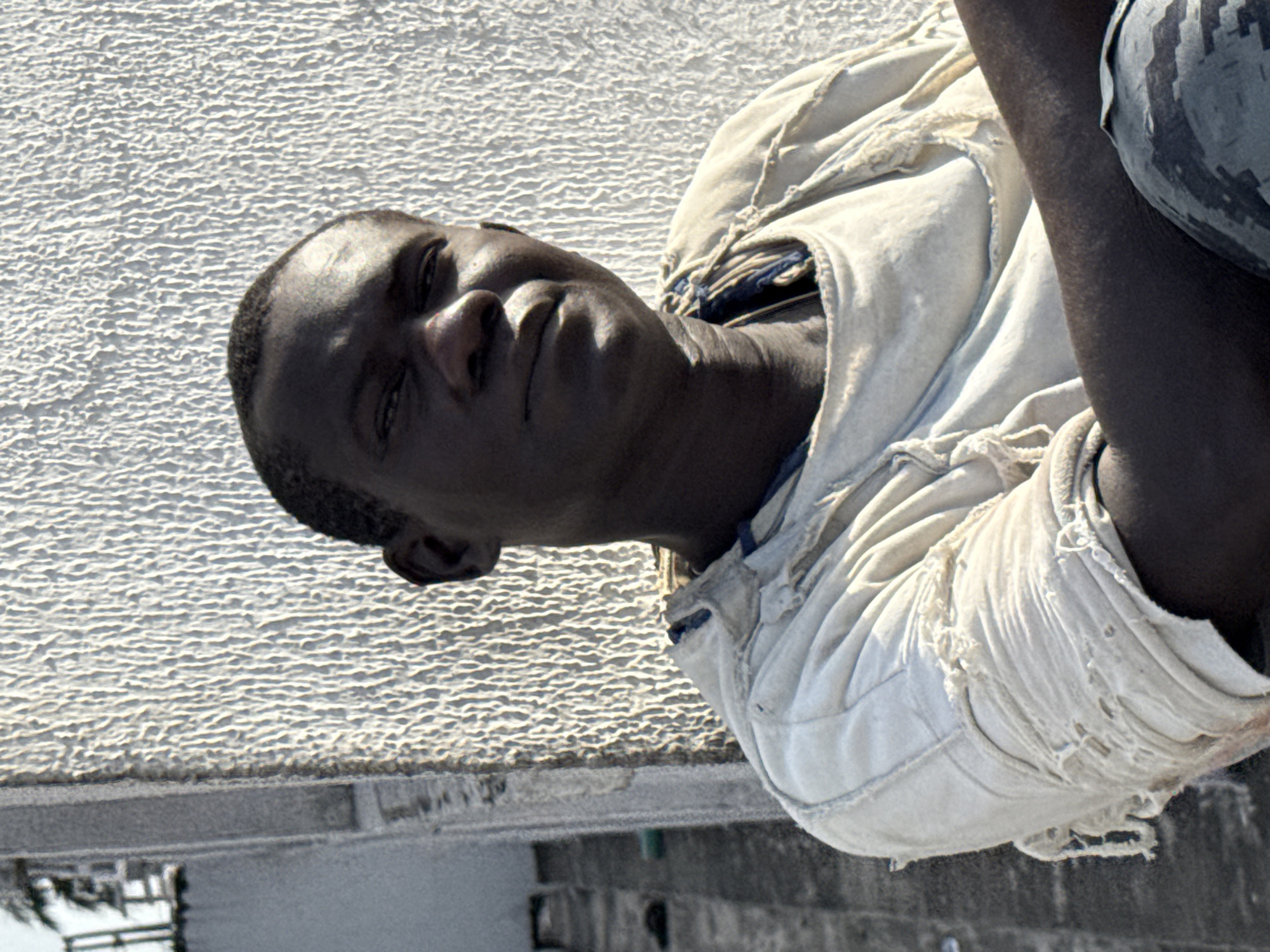 A low-angle medium shot features a dark-skinned man wearing a light-colored, tattered shirt with what appears to be a camouflage-patterned sleeve. He is looking towards the camera with a neutral expression. The background consists of a textured, light-colored wall. Below the wall, there is a street scene, suggesting an urban environment. The presence of buildings and architectural elements suggests the location may be in the city of Ado, Nigeria.