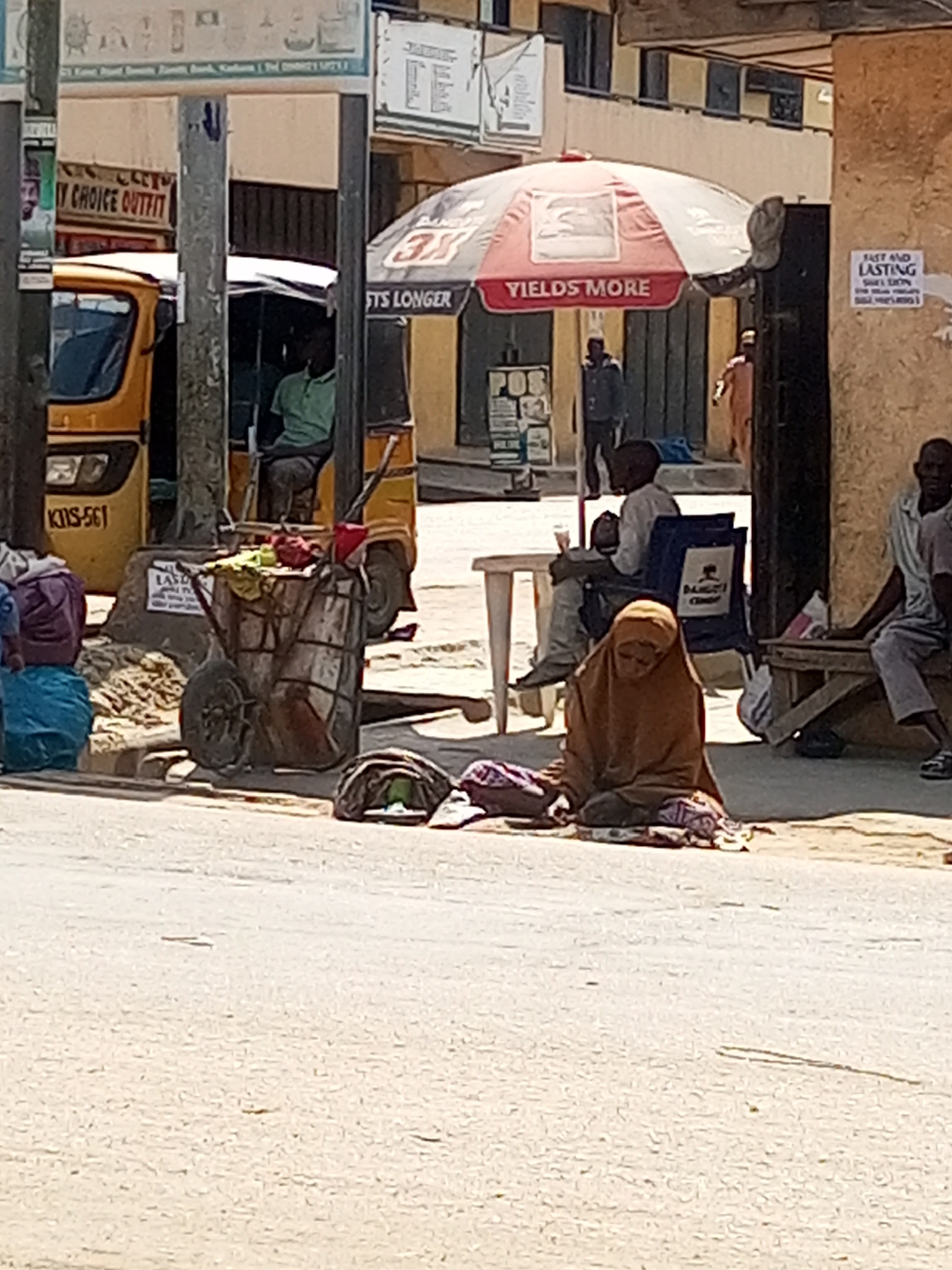 The image depicts an outdoor scene in Maiburiji, Nigeria. A woman wearing a brown hijab sits on the ground, possibly begging. Behind her, a man sits in a blue chair under a red and white umbrella with the text "YIELDS MORE" and "FTS LONGER." Another man sits on a wooden bench against a wall with a sign that reads "FAST AND LASTING." A yellow three-wheeled vehicle, with the license plate "KIS-561," is parked nearby. A cart containing various items stands between the vehicle and the seated woman. Other people are visible in the background. The environment appears to be a street with surrounding buildings.