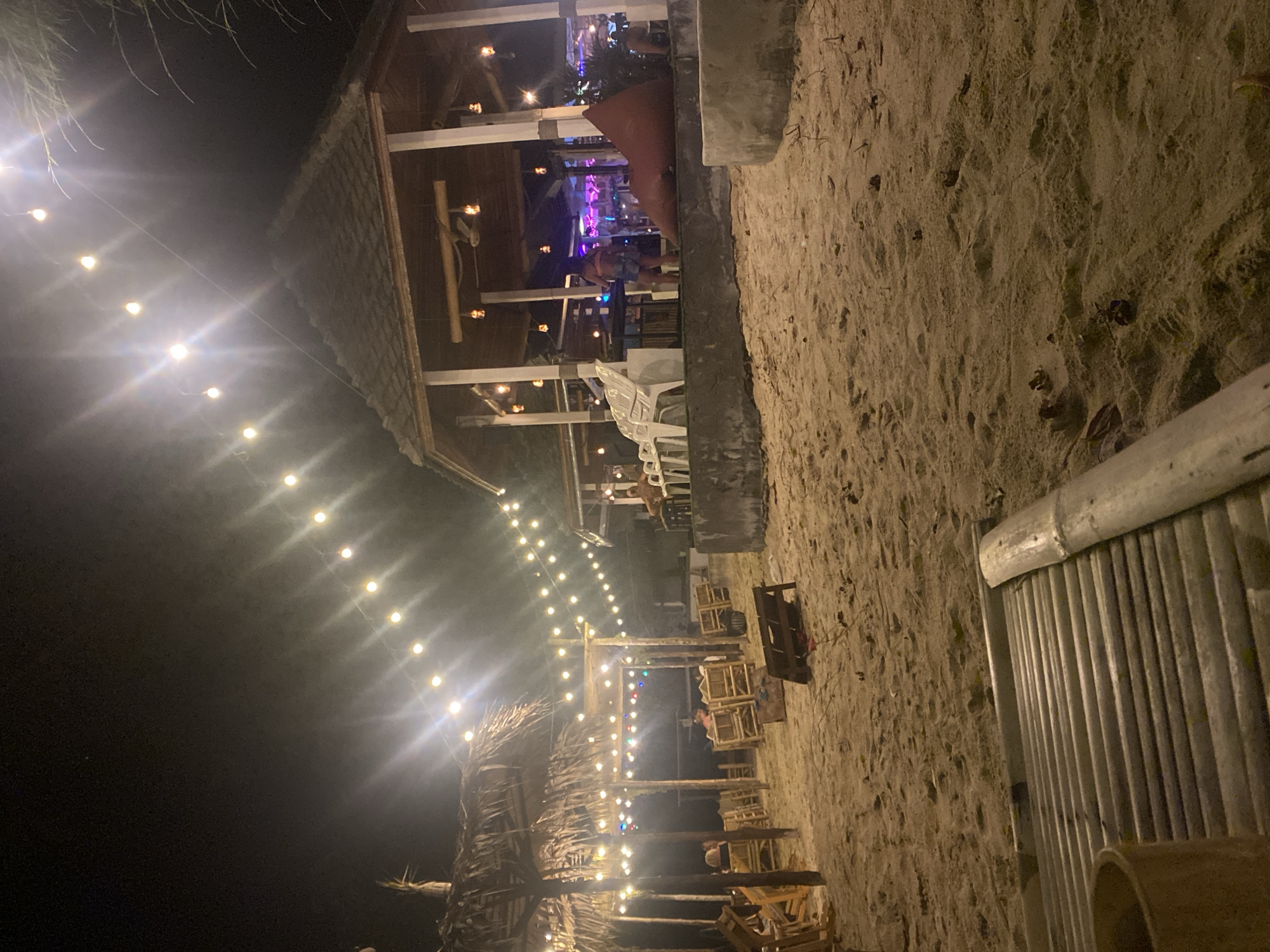 A night scene in Baan Tai, Thailand, shows a beach with outdoor seating under string lights. A structure with a thatched roof is on the left, and a wooden building with open sides is in the background. Tables and chairs are arranged on the sand. People are visible within the lighted building. A section of bamboo fencing is visible on the right side of the frame.