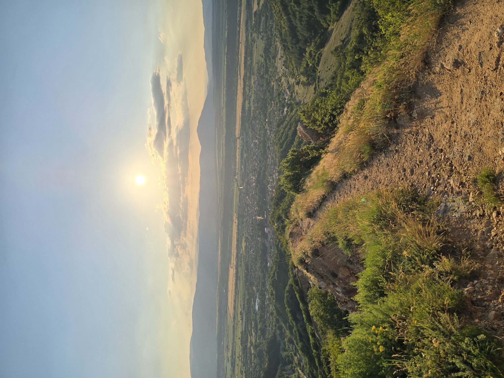 The image displays a panoramic landscape captured from a high vantage point near the city of Stob, Bulgaria, at sunset. In the immediate foreground, a well-defined dirt trail, composed of reddish-brown soil and small rocks, curves gently from the lower left towards the mid-right. The path is flanked by patches of dry, golden-brown grasses and various green shrubs, some exhibiting small yellow blossoms. The terrain on the steep slopes adjacent to the path appears eroded and rocky. In the midground, the valley floor below reveals the clustered buildings of the city of Stob, interspersed with green trees and cultivated fields. Beyond the city, a mosaic of agricultural fields, ranging from dark green to lighter golden-brown, stretches across the valley, enclosed by a distant horizon line. Multiple layers of hazy, undulating mountain ranges are visible in the background. The sky above is characterized by a setting sun positioned in the upper-center, emitting a bright golden light and casting a warm glow across scattered clouds that show both illuminated edges and darker undersides. The upper portion of the sky is a clear, pale blue, transitioning to warmer tones near the horizon. No individuals or specific actions are discernible within the scene.