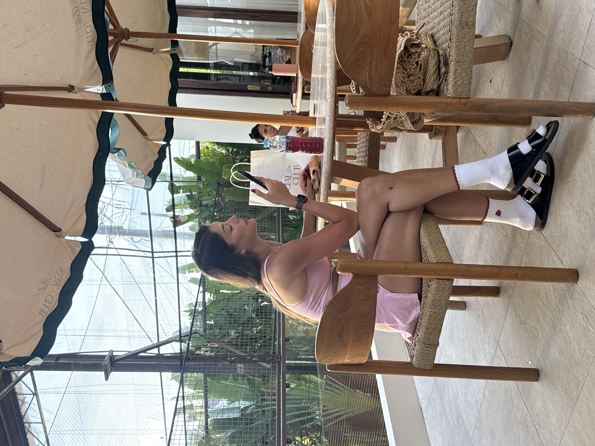 The image shows a woman sitting at a table in North Kuta, Indonesia, based on the "SOCIAL CLUB" printed on a bag next to her. She is fair-skinned, wearing a light pink outfit, white socks with red trim and a small red strawberry design, and black sandals with gold buckles. She is looking at her phone and wearing a watch. She is sitting in a wooden chair, with her legs crossed at the knees. There's an umbrella overhead. The table is round and wooden, with another person sitting at it in the background. There's a bag on the table, and the background shows a garden or green space.