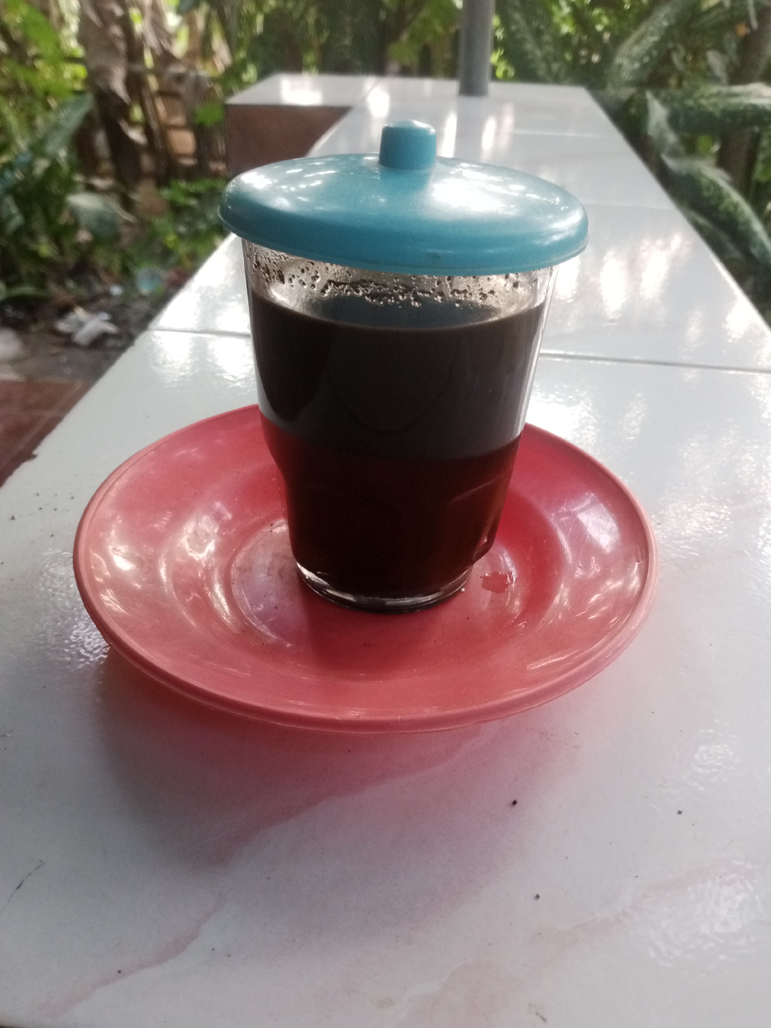 The image shows a close-up view of a glass of dark liquid, possibly coffee, topped with a light blue lid. The glass sits on a pink saucer, which rests on a white tiled surface. The background is blurred, with hints of green foliage and wooden structures, suggesting an outdoor or semi-outdoor setting. The lighting appears natural, casting subtle shadows on the table. The overall impression is one of a casual, everyday scene. The image was captured in Karangpakel, Indonesia.