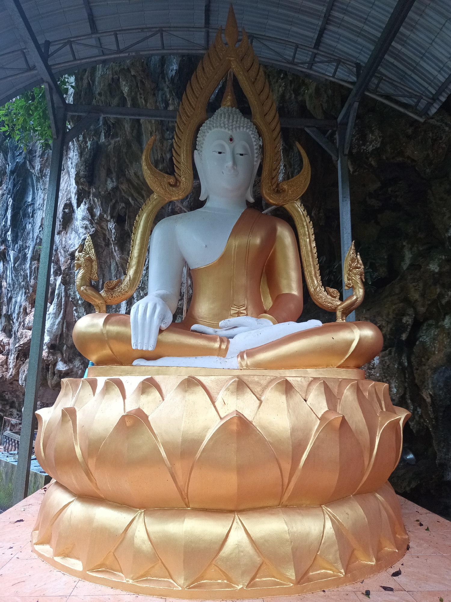 A full-length, eye-level shot depicts a golden and white Buddha statue in an outdoor setting. The statue is seated on a golden lotus flower pedestal with its legs crossed, the right hand placed on the left. The statue’s body is gold, while the head and hands are white. Behind the statue is a rock wall. A partial metal roof is visible above the statue. The statue appears to be located on a tiled surface. This picture was taken in the city of Unknown, Unknown.