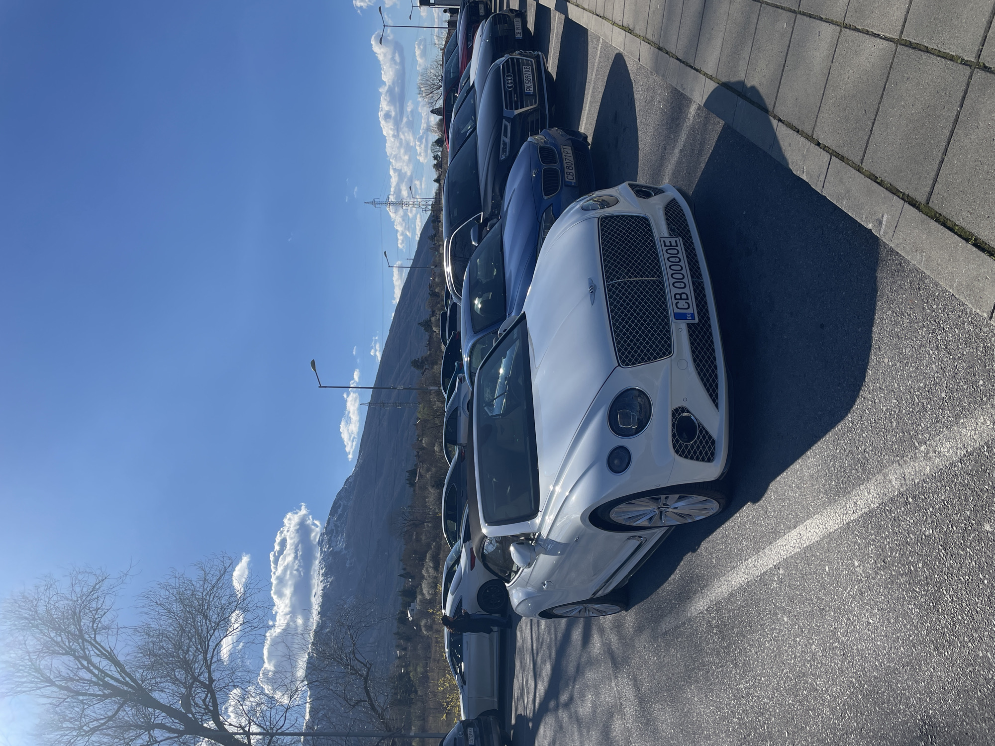 The image shows a line of parked cars on an asphalt parking lot in Sofia, Bulgaria, under a clear blue sky. A white Bentley is in the foreground with license plate "CB 00000E". Parked behind it are several other luxury cars including a blue BMW with license plate "CB 8071PT" and a dark grey Audi with license plate "PX 5097 KB". To the side of the parking lot is a paved walkway and in the background is a mountain range. Sparse, leafless trees are visible along the edge of the parking lot. Street lights are present. The image appears to have been taken during the daytime.