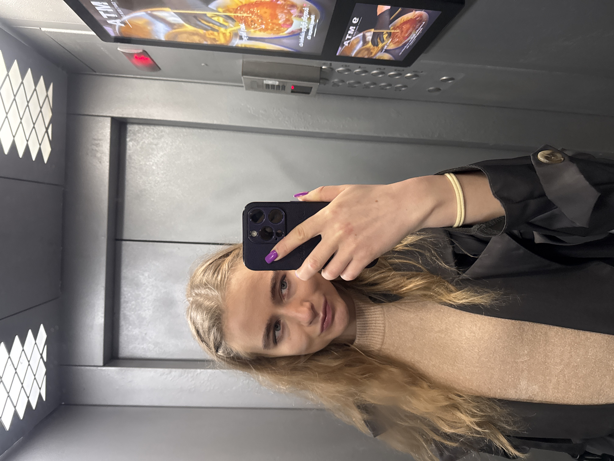 A blonde woman takes a selfie in an elevator. She's wearing a tan sweater and a black jacket with a visible button. She is holding a dark-colored smartphone with a purple case. Her nails are painted purple. The elevator walls are gray with patterned vents on the left. An informational screen and button panel are visible above.