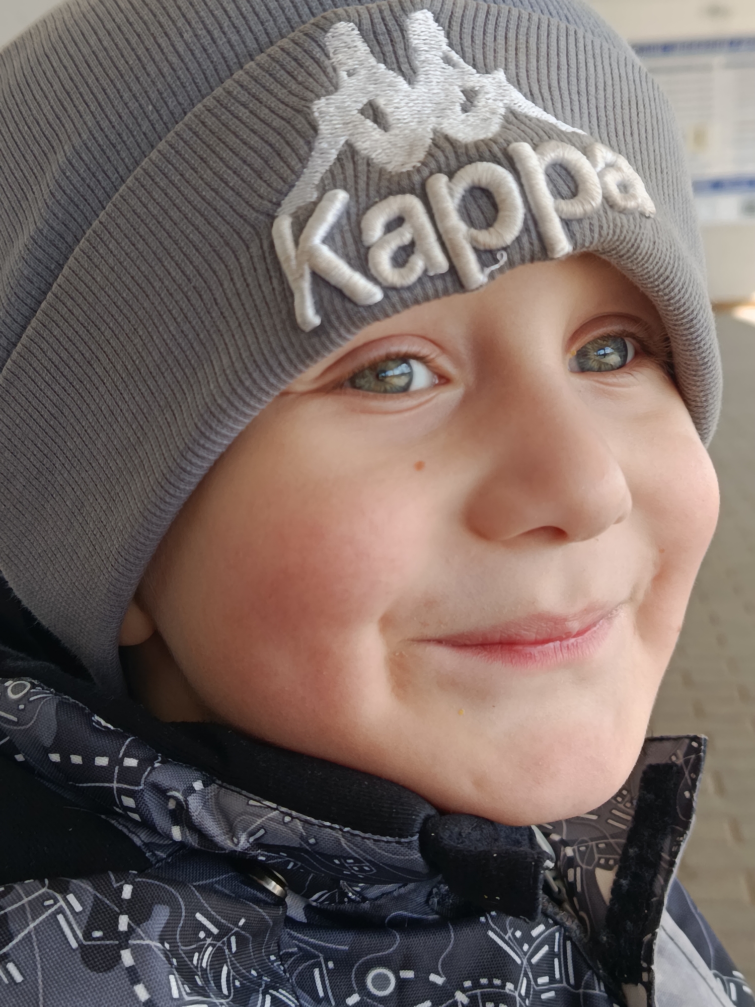 A close-up shot depicts a young child wearing a grey Kappa beanie and a black and grey patterned jacket. The child is smiling, with pink cheeks and visible freckles. The eyes are a light green color. The background appears to be an outdoor urban setting, possibly a city street or building exterior, with a pale brick wall visible. The location is Unknown, Unknown.