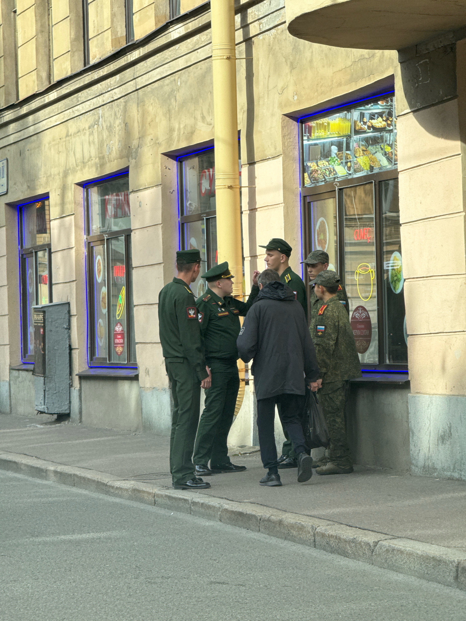 A group of five individuals, four in military uniforms and one in civilian attire, are gathered on a sidewalk in front of a building that appears to be a small food establishment. The building has a light-colored facade and several windows displaying food items and illuminated signage. The street in front of the building is paved and has a concrete curb.
The individuals in uniform are wearing dark green outfits, some with visible insignia on their sleeves. Three of them are facing towards the civilian, who is wearing a dark jacket and pants and is carrying a bag. The fourth uniformed individual is standing slightly apart, facing the building. The interaction appears to be a conversation or a brief meeting.
The environment suggests an urban setting, possibly in Saint Petersburg, Russia, given the architectural style of the building and the type of signage visible in the windows, which includes Cyrillic script. The presence of a yellow utility pole and a gray metal box on the sidewalk further contribute to the urban context. The lighting indicates it is daytime, with sunlight casting shadows.