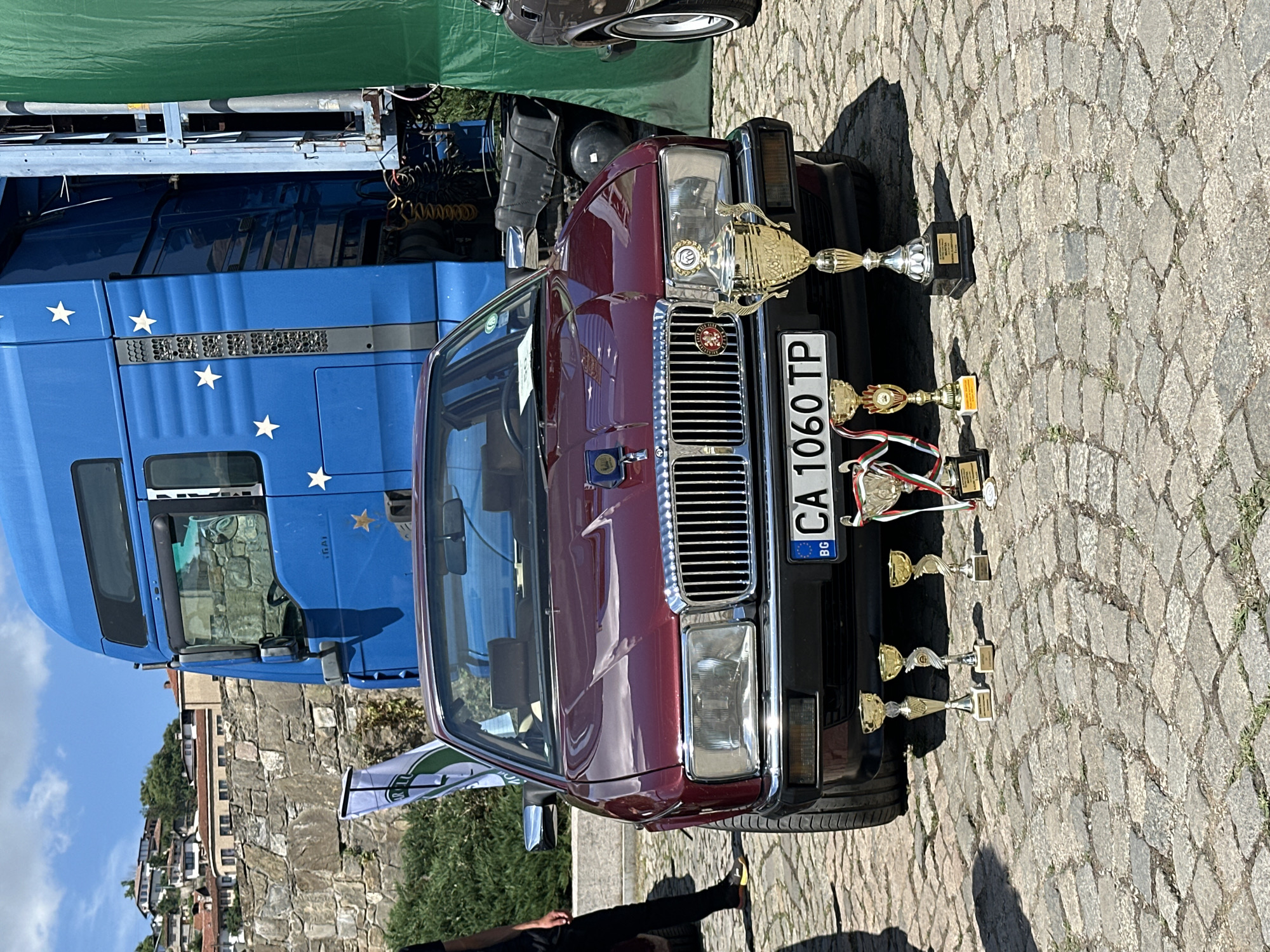 A maroon Jaguar XJ sedan is parked on a cobblestone street in Veliko Tarnovo, Bulgaria, in front of a large blue truck. The car's license plate reads "BG CA 1060 TP". Several trophies are displayed on the ground in front of the car. In the background, stone walls and traditional houses are visible on a hill. A green tarp hangs to the right of the truck. The sky is blue with scattered clouds.