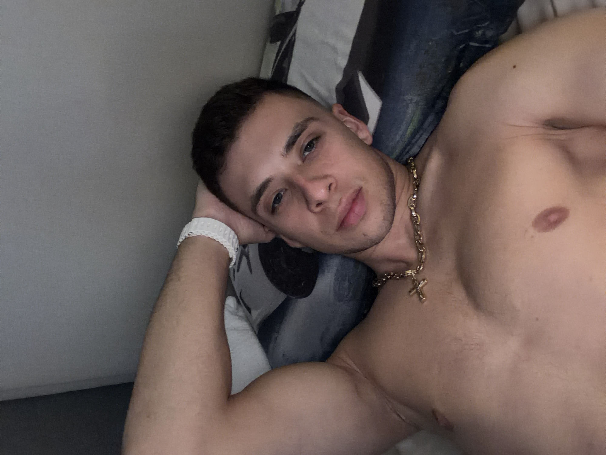 A shirtless man with short dark hair is lying on a bed, resting his left arm under his head. He is wearing a white fitness tracker on his left wrist and a gold chain necklace with a cross pendant. The background includes a white pillow with gray patterns and a light gray wall. The location is not discernible from the image and cannot be determined to be in Plovdiv, Bulgaria.