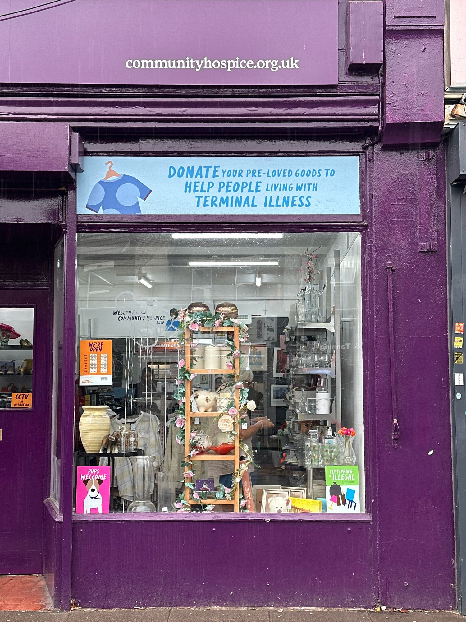 The image shows the exterior of a charity shop with a prominent purple facade. Above the shop window, a sign reads "communityhospice.org.uk". Below this, a blue banner displays text in white: "DONATE YOUR PRE-LOVED GOODS TO HELP PEOPLE LIVING WITH TERMINAL ILLNESS".
The shop window reveals a display of various items, including clothing, vases, decorative objects, and shelves stocked with what appear to be homewares and trinkets. One shelf prominently features ceramic pig figurines and a red bowl. A wooden stand decorated with artificial flowers and greenery is also visible.
On the left side of the window, a "WE'RE OPEN" sign with opening hours is displayed. A pink sign featuring a dog reads "PUPS WELCOME". On the right side of the window, a green sign states "FLYTIPPING IS ILLEGAL". A small CCTV sticker is also visible on the purple door to the left.
The shop is situated on a street, with a pavement in the foreground. The overall impression is that of a retail establishment focused on charity donations. The image is taken from a frontal perspective, with no identifiable landmarks to specify the exact location within London, United Kingdom. There are no people clearly visible in the image, though hands are visible through the glass, suggesting someone might be inside the shop.