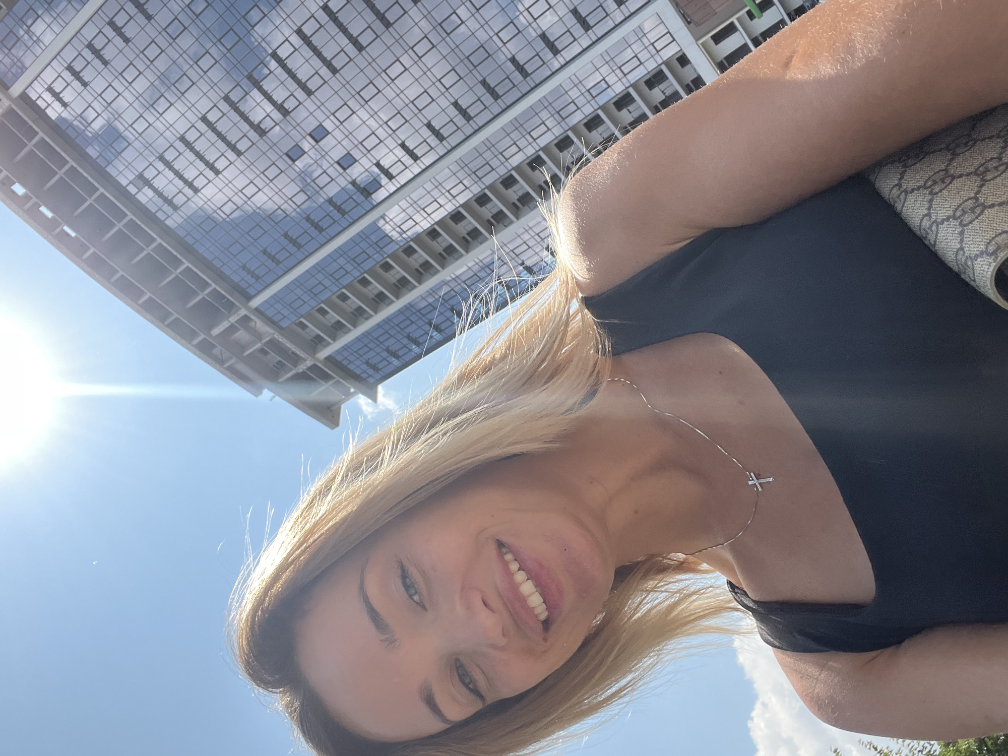 An outdoor, close-up, low-angle selfie shows a blonde woman smiling at the camera. She is wearing a black top and a silver necklace with a small cross pendant. The woman's blonde hair is blowing in the wind. Behind her, a modern glass-fronted building under construction rises against a bright blue sky with the sun prominently displayed. The image appears to have been taken in Fontanka, Ukraine, due to the distinct architectural style of the building and the presence of a canal, which is characteristic of the area.