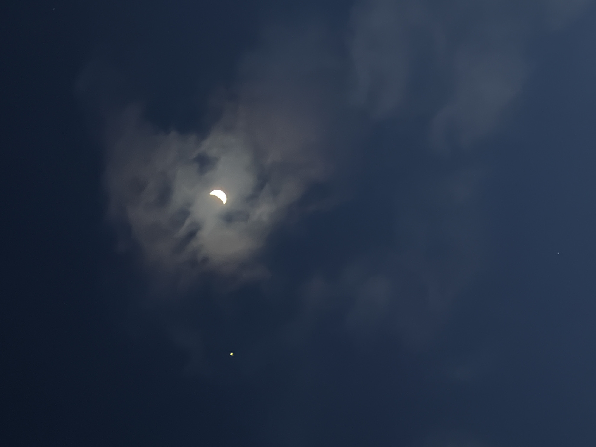 The image captures a celestial event in a dark blue, cloudy sky. The central subject is a partial solar eclipse, where the sun appears as a brilliant white-yellow crescent, peeking through a dense formation of grey and white clouds. The distinct, dark circular edge partially obscuring the sun indicates the moon's silhouette during the eclipse. A soft, bright glow emanates from behind the clouds around the visible crescent.
The sky, though during what appears to be daytime due to the eclipse, is rendered in deep indigo and dark blue hues, typical of an eclipse's effect on ambient light. Significant cloud cover is present, with a dense, textured mass of clouds surrounding the eclipsed sun, and lighter, wispy clouds scattered across the frame.
In addition to the eclipse, two other celestial bodies are visible. A small, faint white point, likely a star or planet, is present in the upper left quadrant. More prominently, a bright, yellowish-white star or planet is visible in the lower central part of the sky, below the eclipsed sun.
The scene depicts the sky over Votkinsk, Russia, during what is likely a solar eclipse, characterized by the dark sky, clouds, and the distinct crescent shape of the partially obscured sun. There is no visible text in the image.