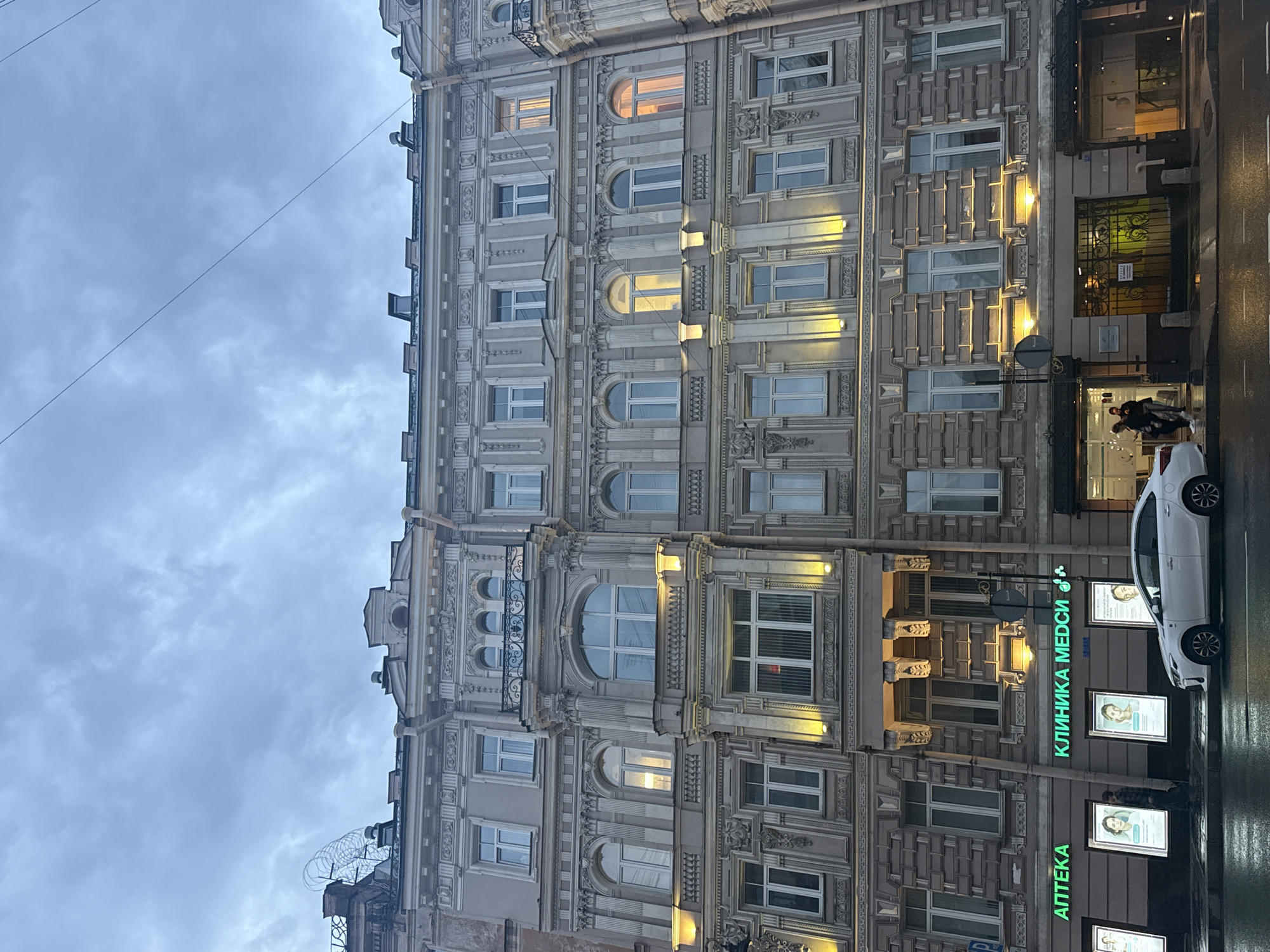 This is a street-level, eye-level, medium shot of an ornate, multi-story building in Saint Petersburg, Russia. The building's facade is a light beige color with intricate architectural details, including columns, decorative moldings, and multiple rows of windows. Many windows on the upper floors are illuminated with warm light, suggesting the time of day is evening or dusk. The sky above is overcast and gray.
On the ground floor, there are commercial establishments. To the left, a green neon sign reads "АПТЕКА" (Apteka), indicating a pharmacy. Next to it, another green neon sign reads "КЛИНИКА МЕДСИ" (Klinika MEDSI), signifying a medical clinic. In front of these establishments, there are illuminated display panels featuring portraits of women.
A white sedan is parked on the wet street in front of the building. Two individuals, dressed in casual attire, are walking on the sidewalk near the entrance of the medical clinic. The street and sidewalk appear wet, suggesting recent rain. The overall scene depicts a typical urban streetscape in Saint Petersburg during the evening.