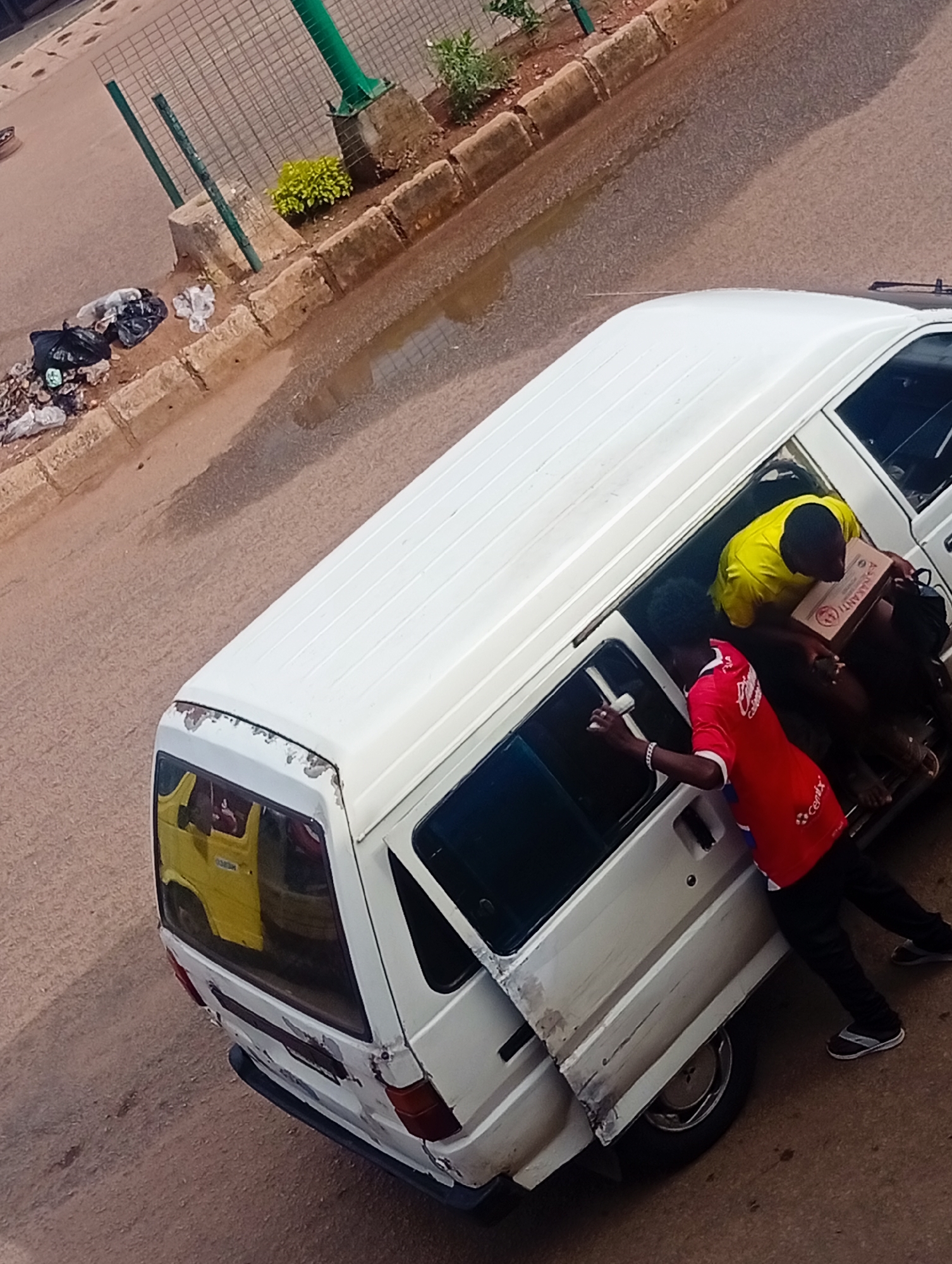 In Bukuru, Nigeria, a white van is parked on a street with a dirt and asphalt surface. The van's rear door is open, and two individuals are interacting with its contents. One person, wearing a yellow t-shirt, is inside the van, holding a cardboard box. The other person, in a red t-shirt, stands beside the open door. Garbage is visible along the curb to the left of the van. A small green plant and a concrete structure are also present on the left side of the image, adjacent to a metal fence. The scene appears to be outdoors, with a wet patch on the road suggesting recent rain.