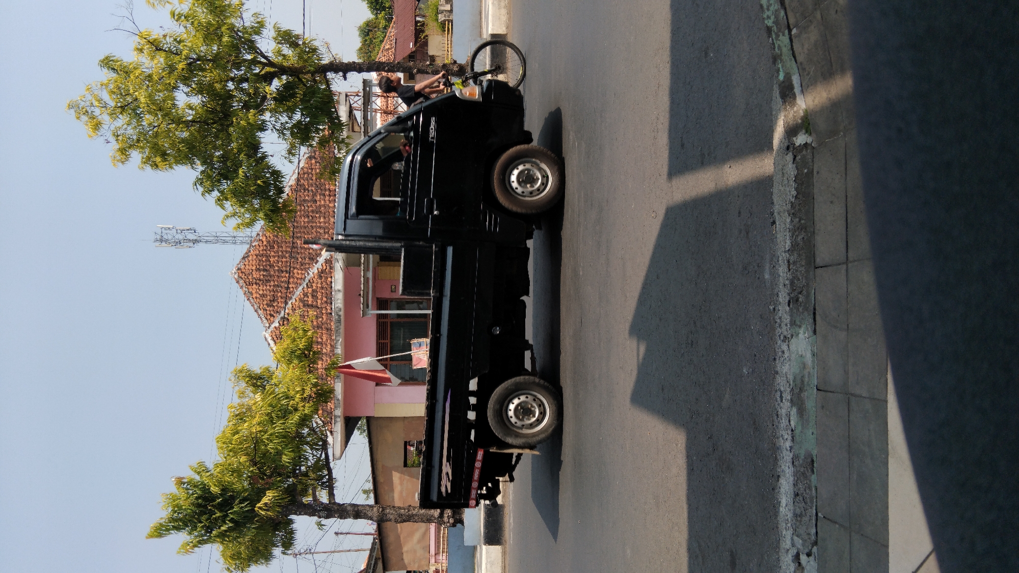 The image captures a street scene in Batang, Indonesia, under a clear, bright sky. A black pickup truck is the central subject, positioned on the asphalt road. To the right of the truck, a person is riding a bicycle, moving in the same direction. Behind the truck, a building with a reddish-tiled roof and a pink facade is visible, with a telecommunications tower rising in the distance. Two trees with green foliage flank the building. The foreground shows the road surface and a paved sidewalk with distinct square tiles, casting shadows from the truck and an unseen object. The scene appears to be an ordinary day with typical urban elements.