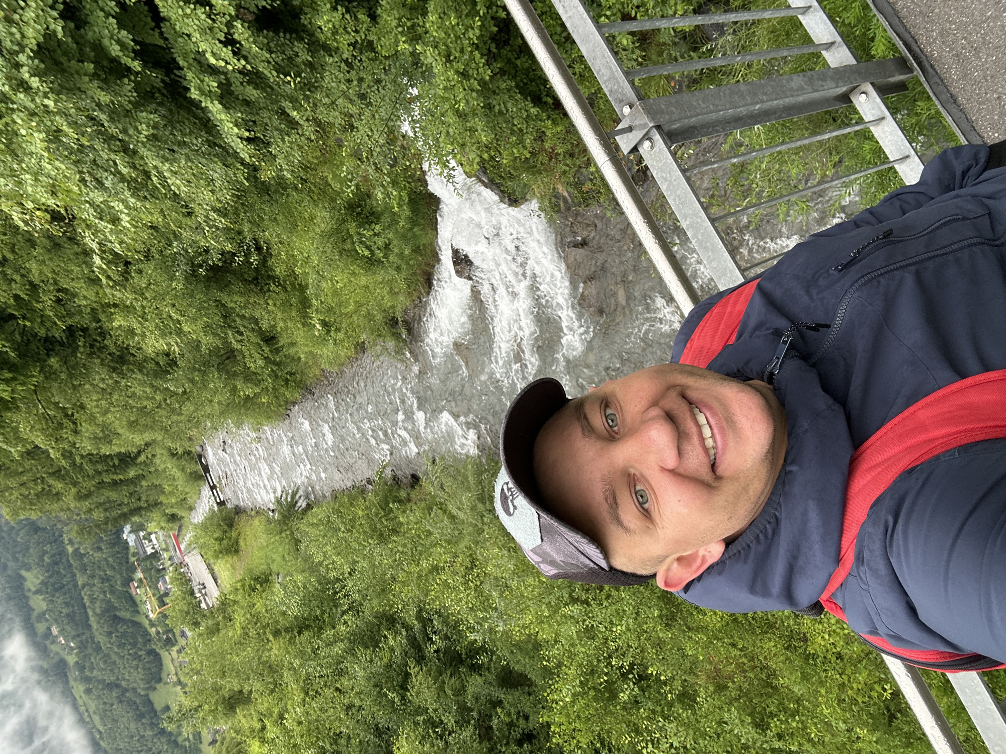 A selfie is taken by a man standing on a bridge with a metal railing. He is wearing a dark blue jacket with a red backpack underneath and a baseball cap. The man is smiling at the camera. Behind him, a river flows rapidly through a lush, green, mountainous landscape. Trees and vegetation line the riverbanks. In the distance, a small town or village is visible nestled in the valley, with buildings and some construction equipment. The sky is overcast, suggesting a cloudy day. The location is the city of Hüsen, Switzerland.