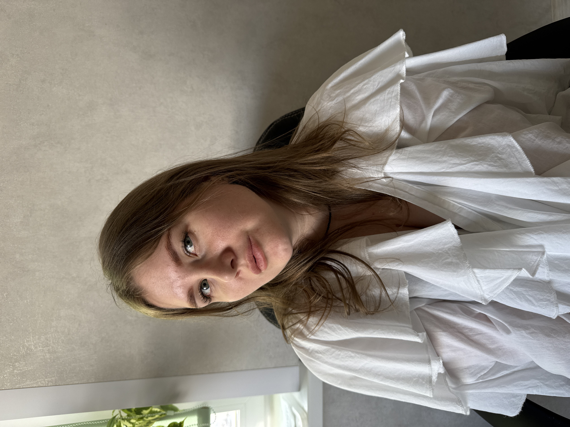 A female individual is depicted in a seated, mid-shot portrait in an indoor setting located in Gornyi (Dvurechenskoe S/P), Russia. The subject has light brown hair, blue-green eyes, and fair skin, with a neutral expression as she looks directly at the viewer. She is wearing a white, ruffled V-neck blouse and a thin dark necklace. The background consists primarily of a light gray textured wall. To the subject's left, a white-framed window and a portion of a green leafy plant are visible. The black backrest of a chair is partially obscured behind the subject. Illumination is consistent with natural light from the visible window combined with internal ambient lighting.