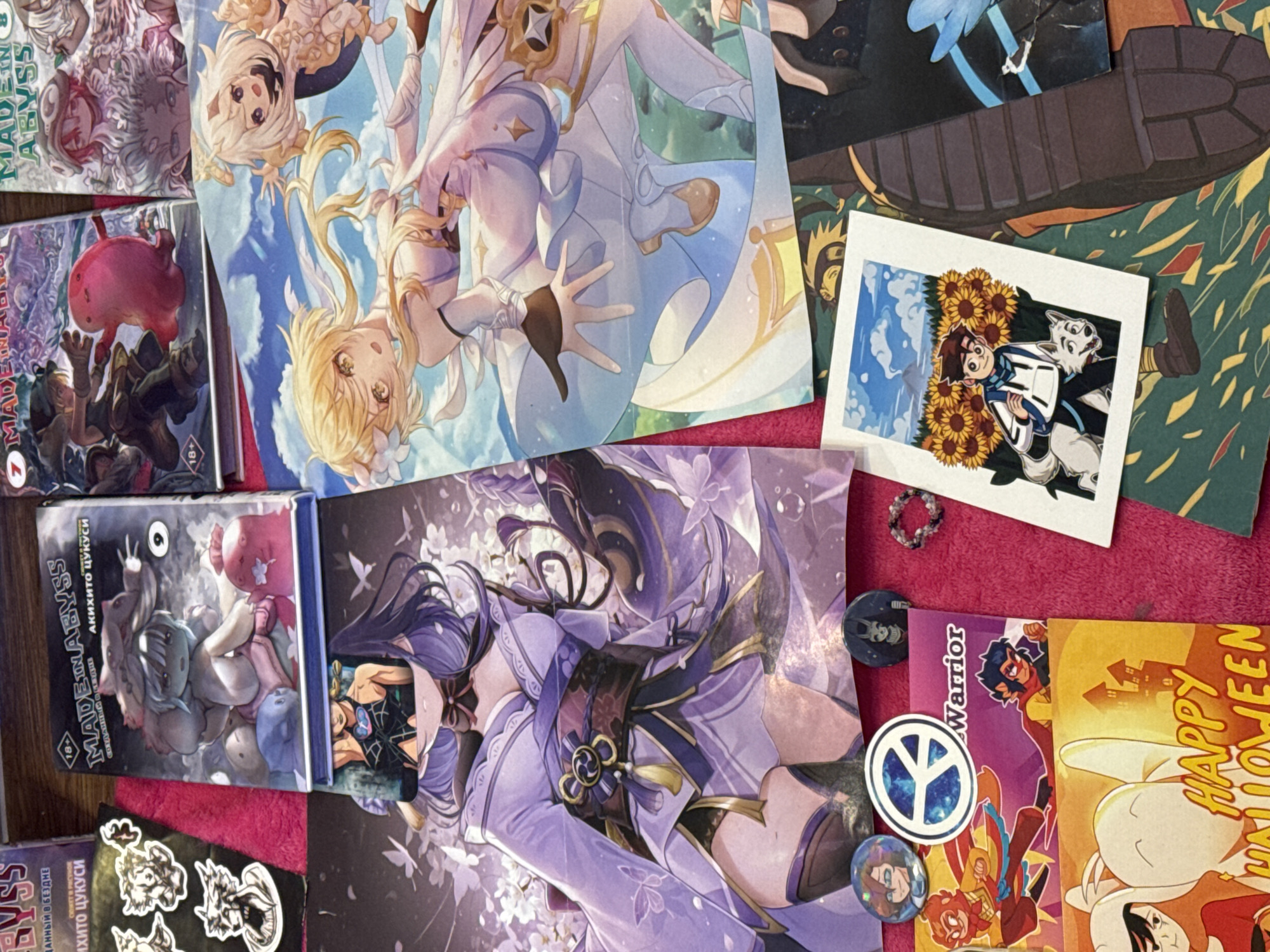 The image presents a flat lay of various anime-themed objects. On the left are several volumes of the manga series "Made in Abyss", with visible Russian text on the spines, indicating a Russian edition. To the right of the manga is a large print featuring characters resembling those from the video game "Genshin Impact." Below this print is another depicting a female anime character with long purple hair, reminiscent of "Raiden Shogun" from the same game.
Scattered around the prints and manga are various smaller items: a polaroid-style print featuring anime characters with sunflowers, several character-themed buttons, a small beaded ring, and other anime-style postcards or prints with characters and text like "Warrior" and "Happy Halloween."
The items are arranged on a surface covered with a bright pink, possibly velvety, cloth. The overall scene suggests a collection of anime and manga merchandise. There is no specific evidence in the image that could definitively link it to Saint Petersburg, Russia.