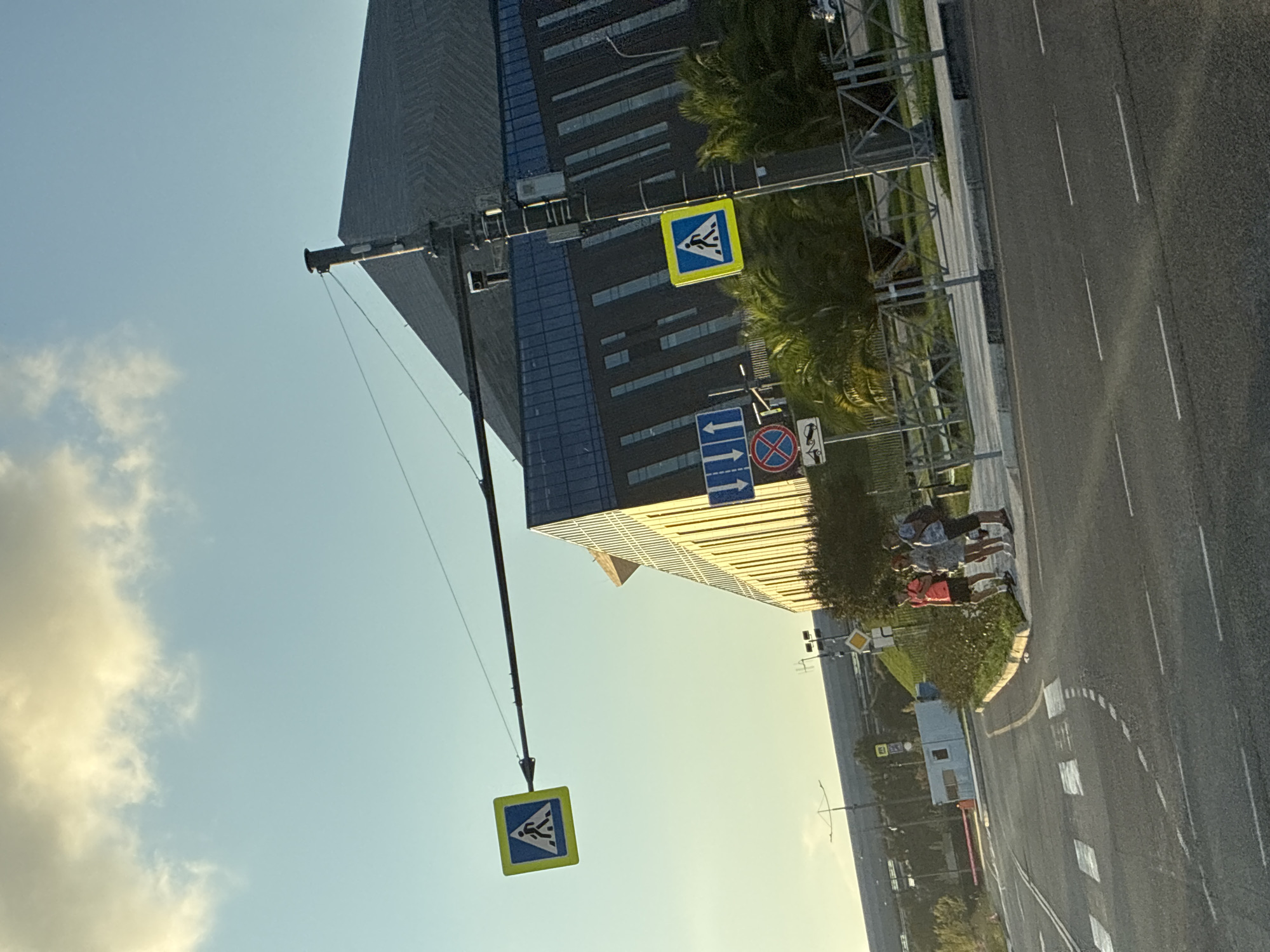 The image shows a street scene in Sirius, Russia, with a modern building in the background. The building has a dark, angled roof and vertical blue and grey panels on its facade. In the foreground, there is a road with pedestrian crossings. Several traffic signs are visible, including two signs indicating a pedestrian crossing, one with a person walking. Other signs include one with three upward arrows and another with a red circle and a diagonal line over a tow truck, indicating no towing.
Three people, two women and one man, are standing on the sidewalk next to the road, appearing to be waiting. They are dressed in casual summer clothing. One woman is wearing a pink top and shorts, another is in a grey dress, and the man is in a blue and white patterned shirt and shorts. They are facing away from the camera, towards the road.
The sky is bright blue with some wispy clouds, and the sunlight casts shadows, suggesting it is daytime. Palm trees and other greenery are visible behind the people and near the building. A light pole with attached equipment and wires is prominently featured, supporting one of the pedestrian crossing signs. The overall environment appears to be an urban or semi-urban area with modern infrastructure.