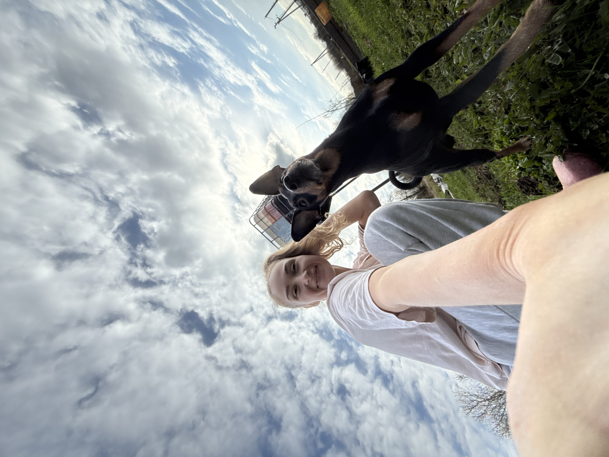 The image is an upward-angled selfie taken outdoors in Nizhnyaya Chermoda, Russia. A young woman with blonde hair is smiling and holding the camera out at arm's length. She is wearing a white t-shirt and grey sweatpants. A black and brown dog stands beside her, its hind legs on the ground and front legs raised in the air. The dog is wearing a muzzle. Behind the woman and dog is a blue sky filled with white clouds. To the right, there is a patch of green grass and some distant trees. A fence and utility pole are visible in the background.