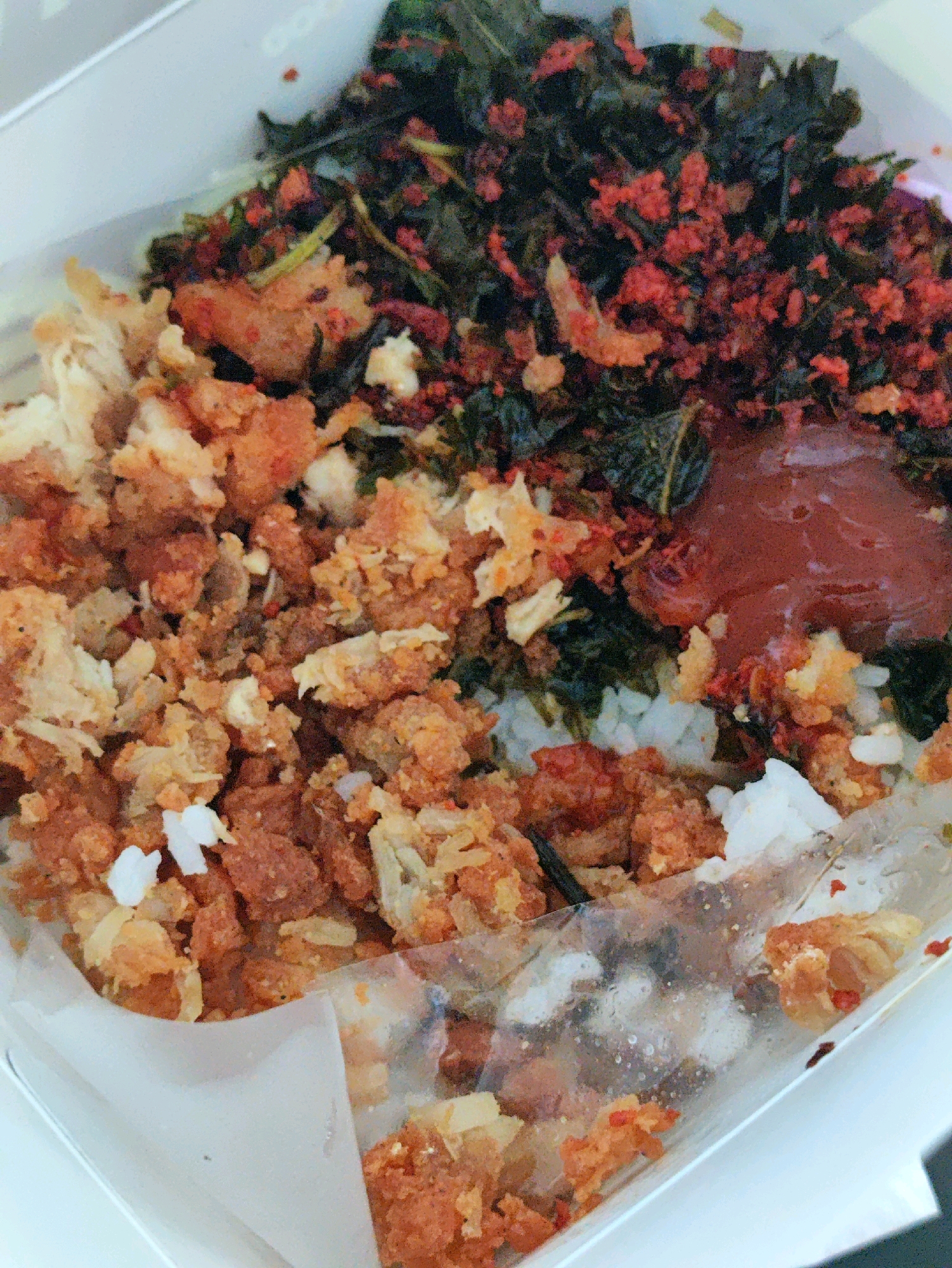 A close-up, overhead shot shows a take-out food container filled with rice, fried chicken, and greens. The food is arranged in distinct sections within a white cardboard container lined with white paper. The rice forms the base, with a generous portion of shredded, golden-brown fried chicken on the left side. To the right of the rice is a pile of dark green leafy vegetables mixed with small, reddish-orange pieces, possibly chili flakes or a spicy seasoning. A dollop of dark red sauce is visible on top of the greens. The container appears to be on a plain, light-colored surface, possibly a table. The image captures the details of the food's texture and color.