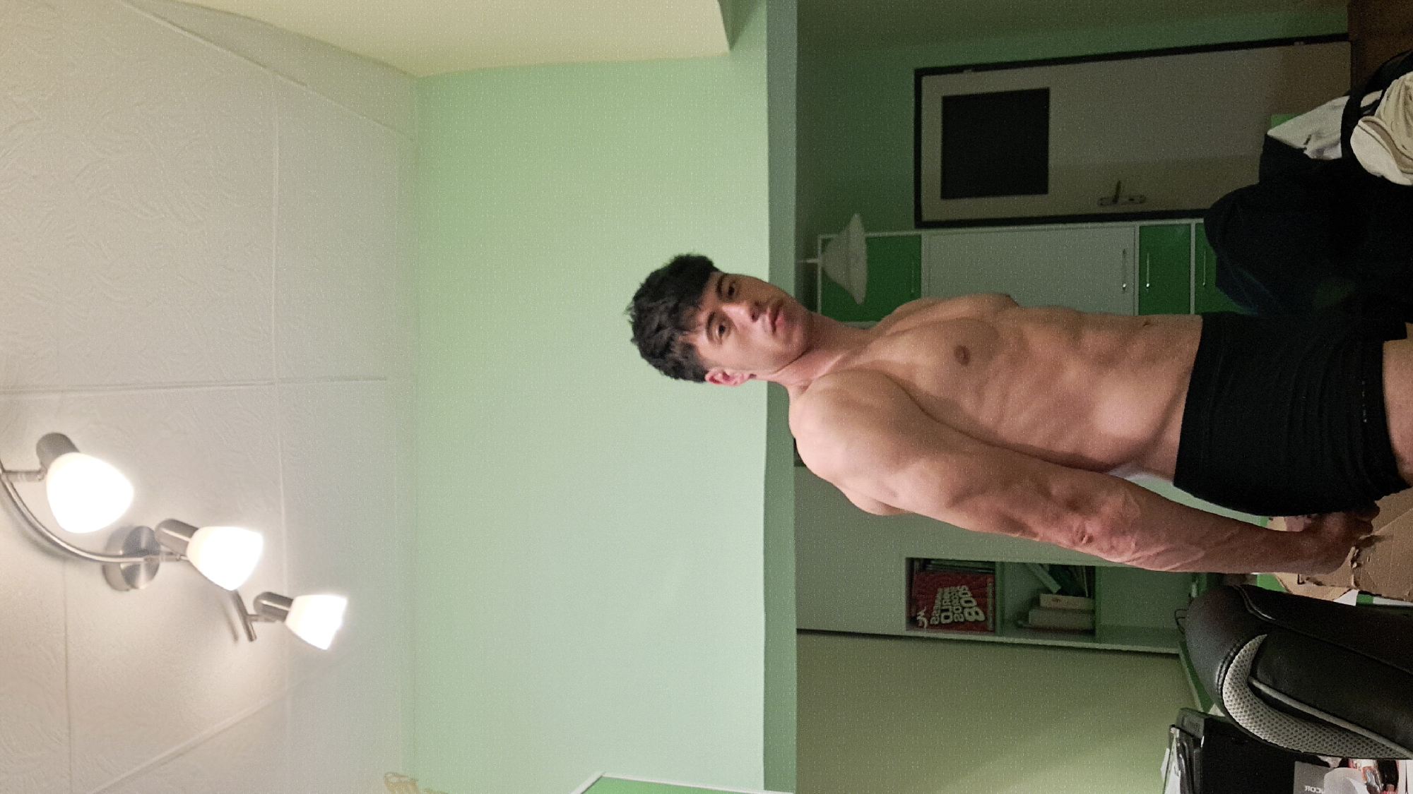 A medium shot depicts a shirtless, light-skinned male with dark, curly hair, wearing black shorts. He is in a room with a pale green wall. A ceiling light fixture with three bulbs is visible on the left. In the background to the right, there is a framed picture on the wall above a white and green cabinet. There is also a black office chair with grey accents in the foreground on the lower right. The city of Burgas, Bulgaria, is not discernible.