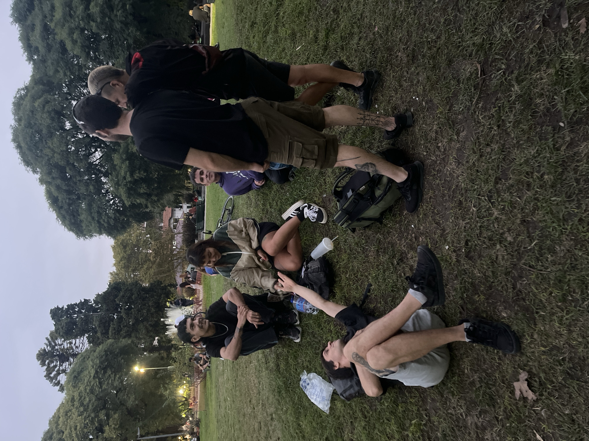 A group of people is on the grass, including two standing males, one with a hat and glasses, the other with a black t-shirt and cargo shorts with tattoos on his legs. Another male in a purple sweatshirt is sitting down. A woman in a tan jacket is sitting near two other males, one in a black shirt, and another lying on his back. There are trees in the background, and light poles. The scene appears to be in a park in the evening. The city appears to be Buenos Aires, Argentina.
