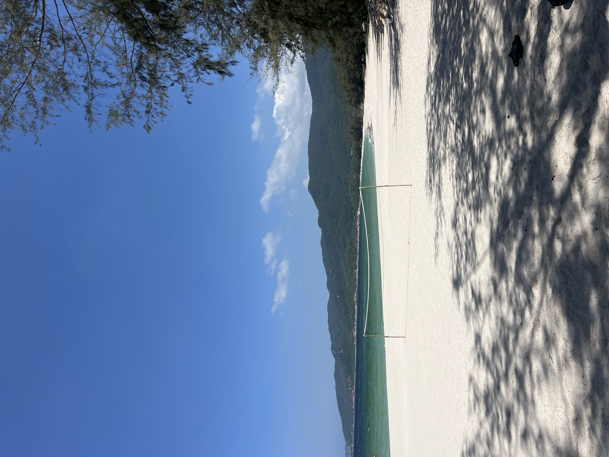 The image displays a sunny beach scene in Cong Hai, Vietnam. A clear blue sky dominates the upper frame, featuring a few small white clouds, primarily on the right side. A range of green, tree-covered mountains forms the distant horizon, separating the sky from the ocean. The ocean water exhibits a gradient, appearing turquoise-green near the shore and transitioning to a deeper blue-green further out. A white volleyball net is set up on the white sandy beach, supported by two wooden posts, positioned close to the water's edge. The wide expanse of white sand is visible, with its foreground heavily patterned by dappled shadows cast by trees located off-frame to the right. Dark green, coniferous trees line the right side of the beach, with some branches extending into the upper right corner of the image. A pair of dark sandals or flip-flops rests on the sand in the lower right, within a shaded area. No human figures are clearly visible on the beach or in the water, though a small, indistinct dark object is present in the water on the far left.