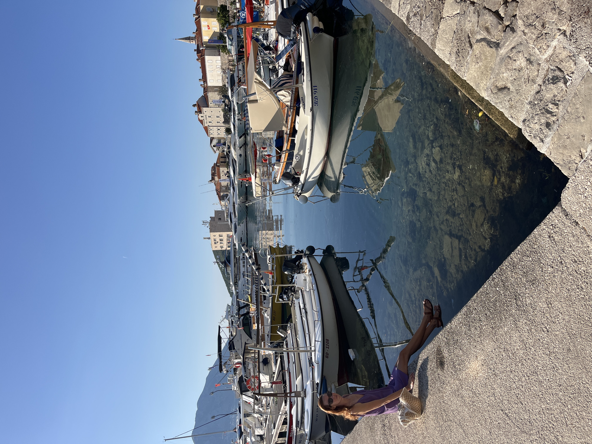 The image captures a marina scene in Budva, Montenegro, under a clear, bright blue sky with a faint aircraft contrail in the upper right. A woman is seated on a concrete pier in the lower left foreground, wearing a purple slip dress, dark sandals, and sunglasses. Her long brown hair is down, and a woven straw bag is positioned next to her left hip. Her legs extend over the water, which is clear enough to reveal rocks and some vegetation on the seabed.
Numerous boats of varying sizes are docked in the marina. On the left, several white and yellow boats are visible, with a prominent white vessel displaying "BD-1108" on its side and another behind it showing "BD 352." Further back on the left, several large yachts are moored. On the right, a white boat with a beige canopy and an outboard motor displays the registration "BD 911" and the name "БИЈЕЛИКА" on its hull. Reflections of boats and the sky are distinctly visible on the water's surface.
The background showcases the architecture of Budva Old Town, featuring light-colored buildings with traditional red-tiled roofs. A prominent church spire is visible on the right side of the Old Town structures. Mountains rise in the distance on the left side of the scene. The pier's surface transitions into a rougher, stone-paved section on the lower right.