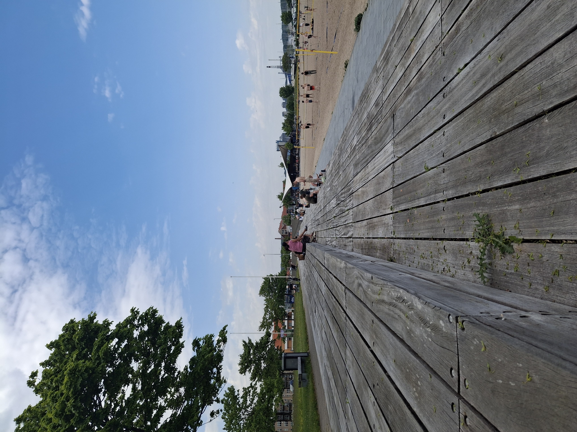 The image depicts a scene on the waterfront in Aabenraa, Denmark, captured on a sunny day with a blue sky and scattered clouds. The primary focus is a weathered wooden boardwalk or deck stretching across the foreground. In the background, a beach area is visible where people are playing beach volleyball and lounging. Further back, there is an urban landscape with buildings and infrastructure. A green tree stands prominently in the lower left corner of the frame. A person is sitting on the edge of the boardwalk.