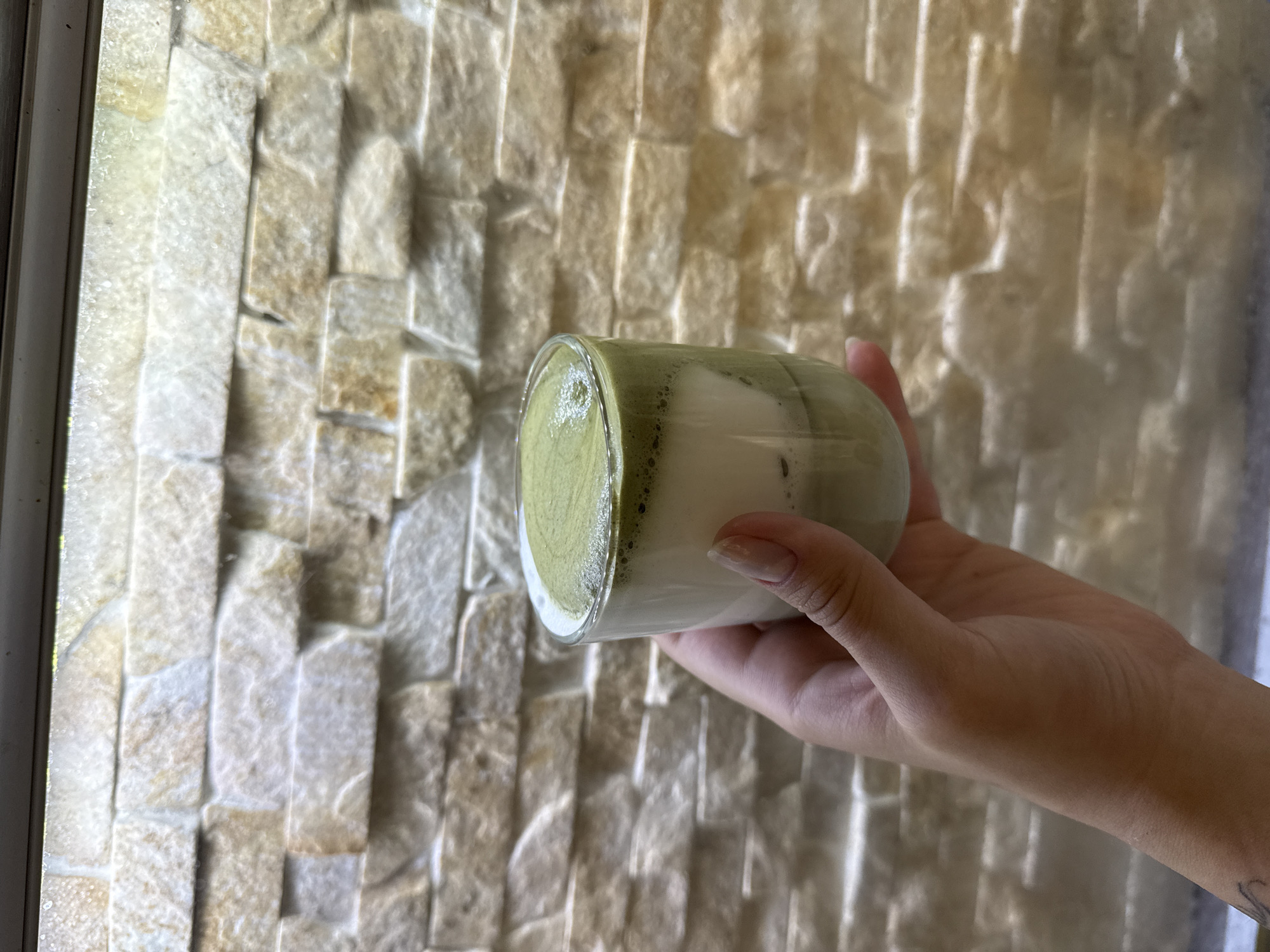 A hand holds a glass containing a layered beverage, likely a matcha latte. The drink has a white layer beneath a frothy green matcha layer. The background is a wall constructed of stacked, light-colored stones, some of which appear to be rough-hewn and others more polished. The lighting suggests an indoor setting, possibly a cafe or restaurant. No specific location within Odesa, Ukraine, can be identified from the visual information provided.