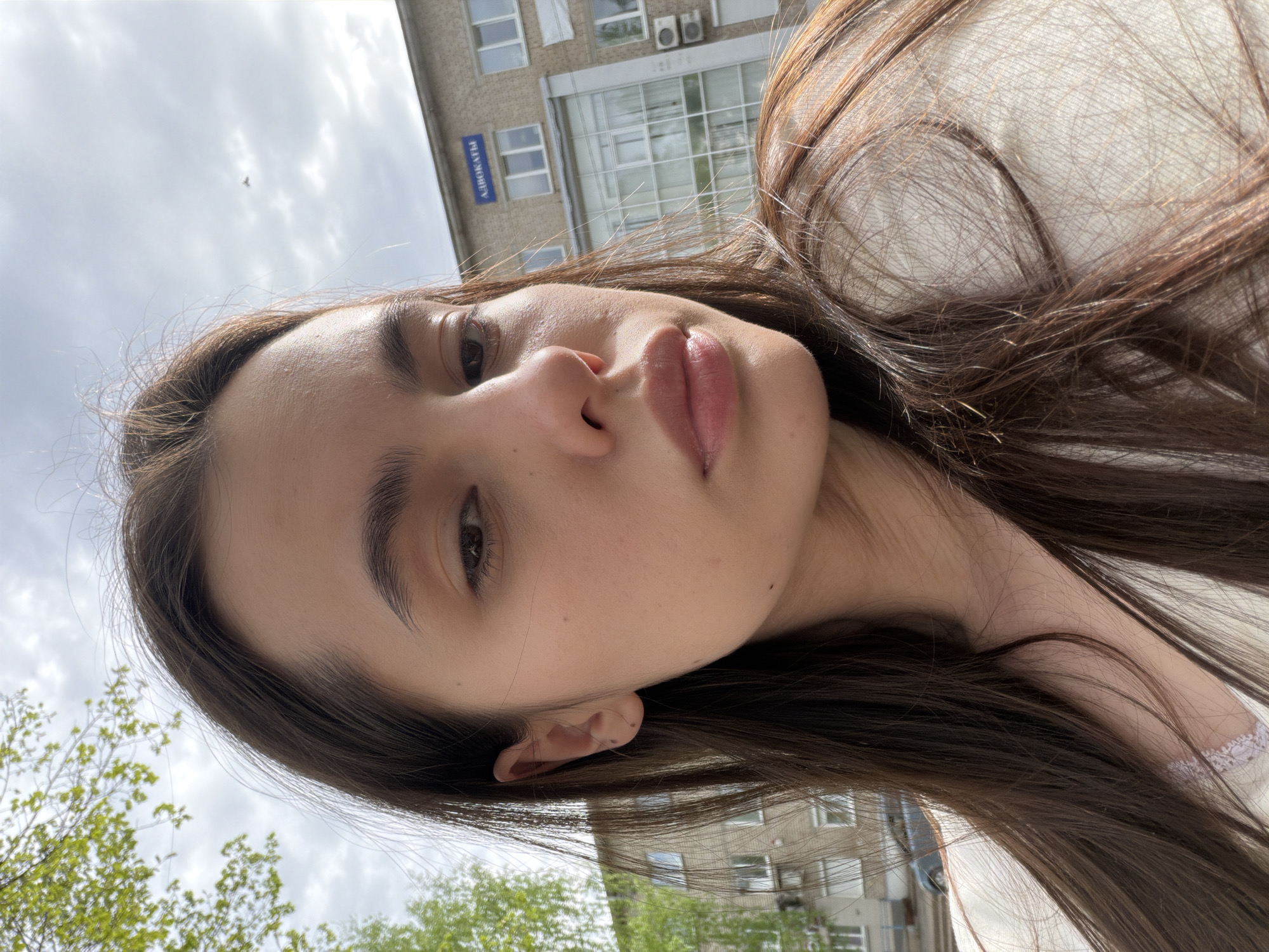 A woman with long brown hair is pictured from a low angle outdoors. Her face is close to the camera, showcasing her features. She wears a white top. In the background, a building is visible. A sign on the building reads "АДВОКАТЫ". The presence of the sign in Russian indicates the location is likely in a Russian-speaking area. Given the query specifies the city of Chaikovsky, Russia, the scene is probably located there. Trees and cloudy sky are also visible.