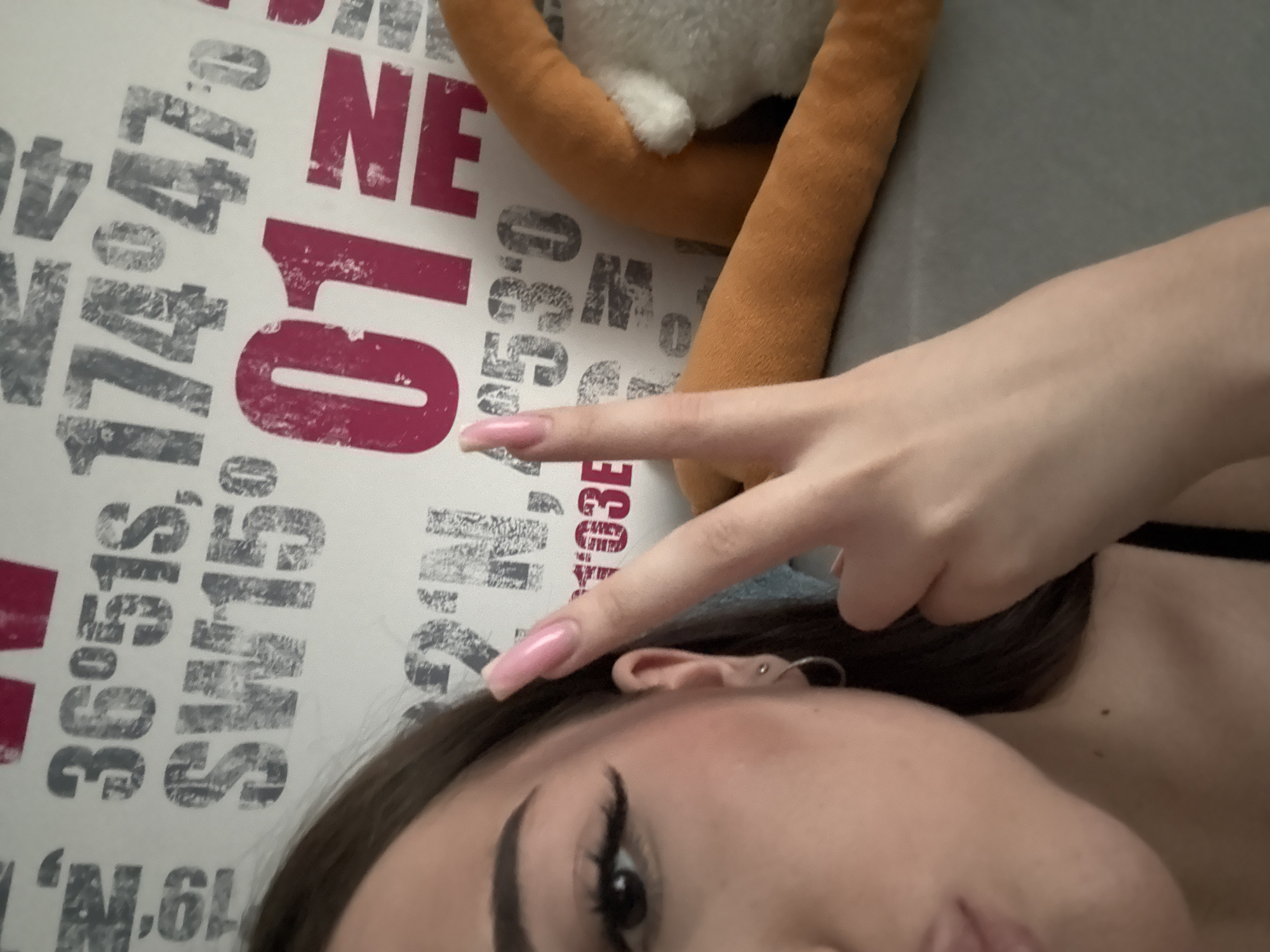 A close-up shot of a person's face, positioned at an angle, with their left hand raised in a peace sign near their ear. The person has dark hair, defined eyebrows, and visible eyeliner. They have an ear piercing with a ring. The background includes a wall with text and numbers printed on it in a distressed style, reading "NE 10" and other coordinates. A portion of an orange and white stuffed animal is also visible in the background. The city of Naberezhnye Chelny, Russia is not evident in the image and cannot be determined based on the visual information provided.
